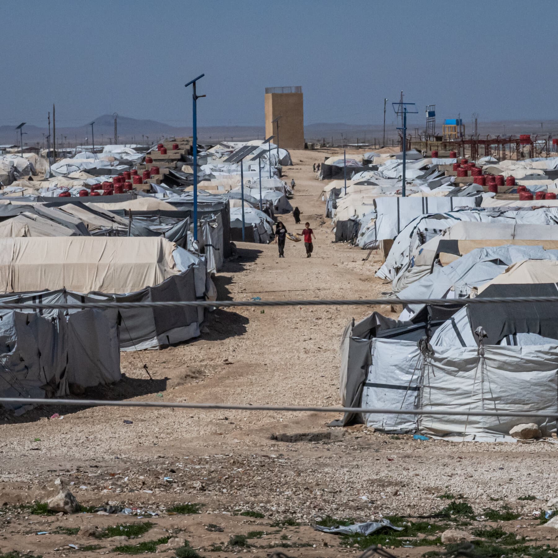 A photorealistic news image of a bored U.S. diplomat shrugging while handing a heavy cardboard box labeled 'TERRORISM & REGIONAL CHAOS' to a confused-looking Syrian military officer across a conference table.