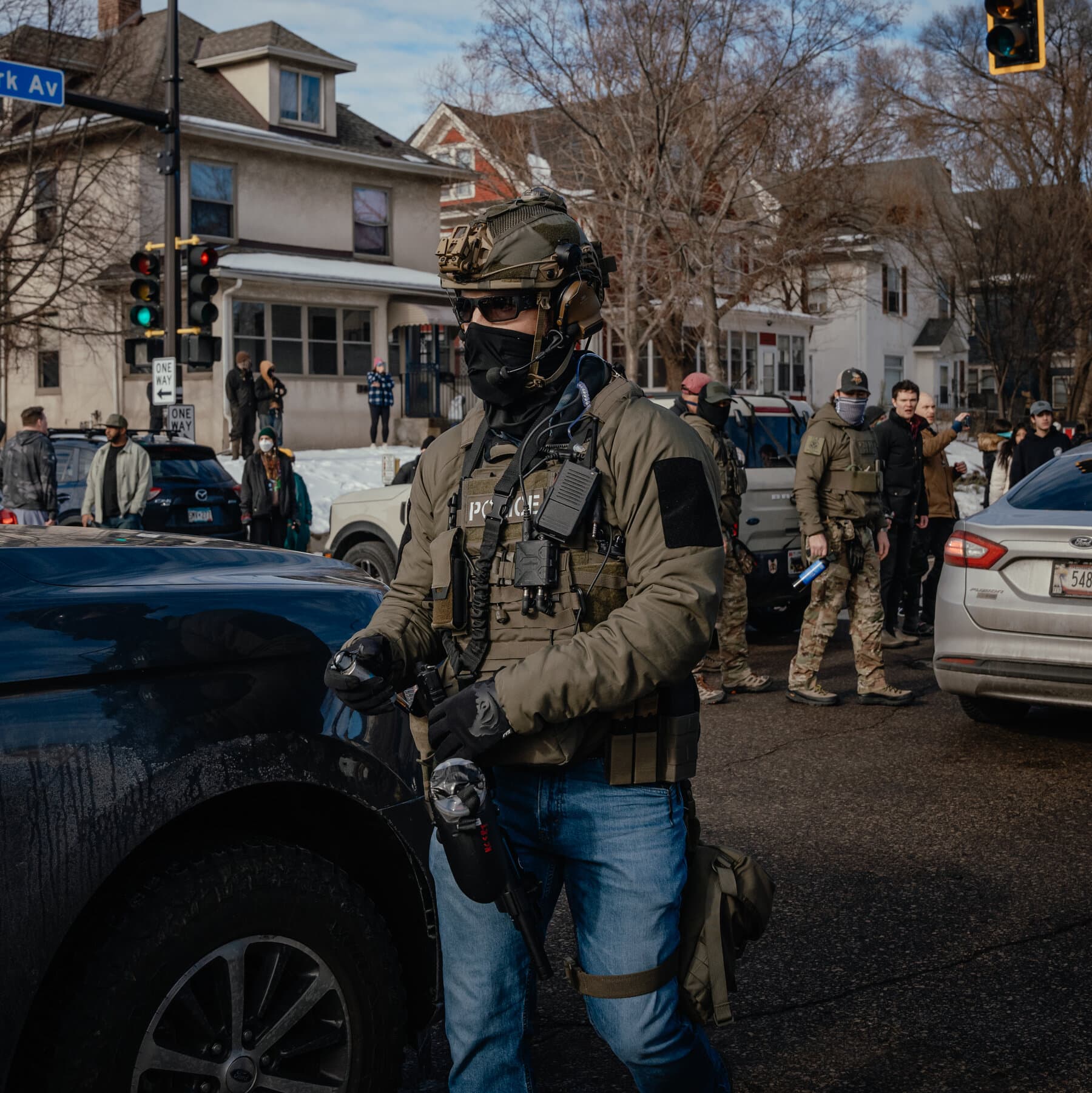 A high-contrast, noir-style political cartoon depicting a snowy Minnesota street. In the foreground, two oversized, faceless bureaucratic figures in tactical gear are pointing fingers at each other. One represents local police with a sheriff star, the other represents federal agents with 'FED' on the vest. They are tangled in messy red tape that is tripping up civilians in the background. The atmosphere is cold, grey, and hostile.