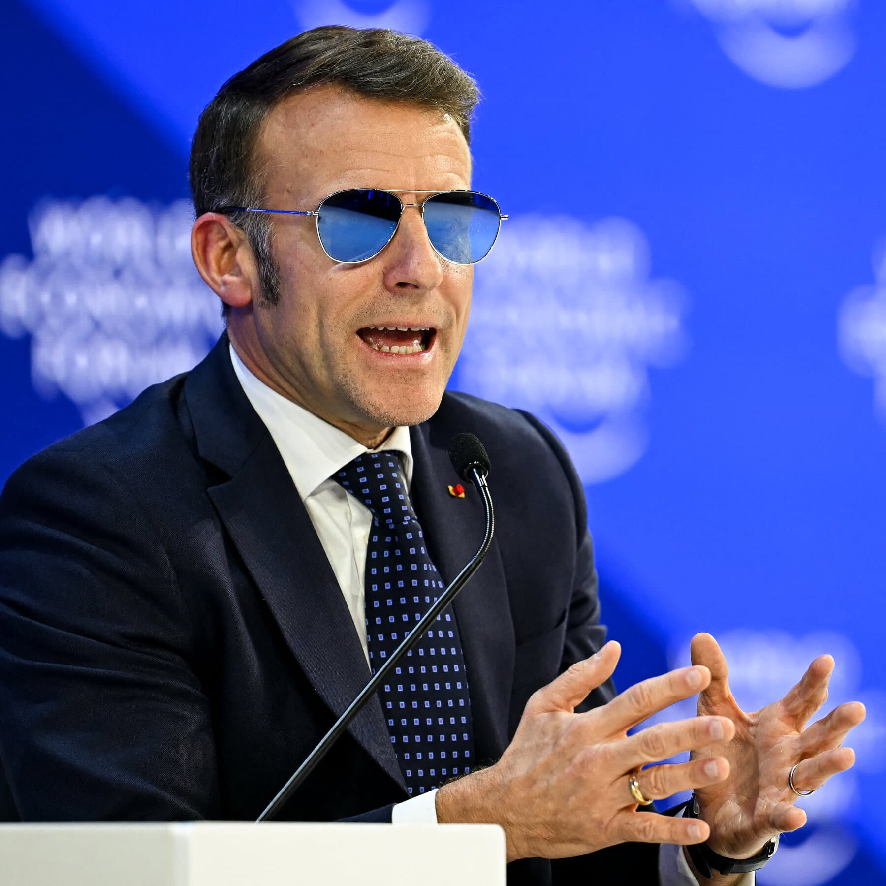 A satirical political cartoon style image depicting a tiny, impeccably dressed Emmanuel Macron standing on a melting glacier, holding a tiny paper shield, screaming at a gigantic, menacing orange storm cloud shaped like a face. The background features the Swiss Alps and confused billionaires in suits drinking champagne.