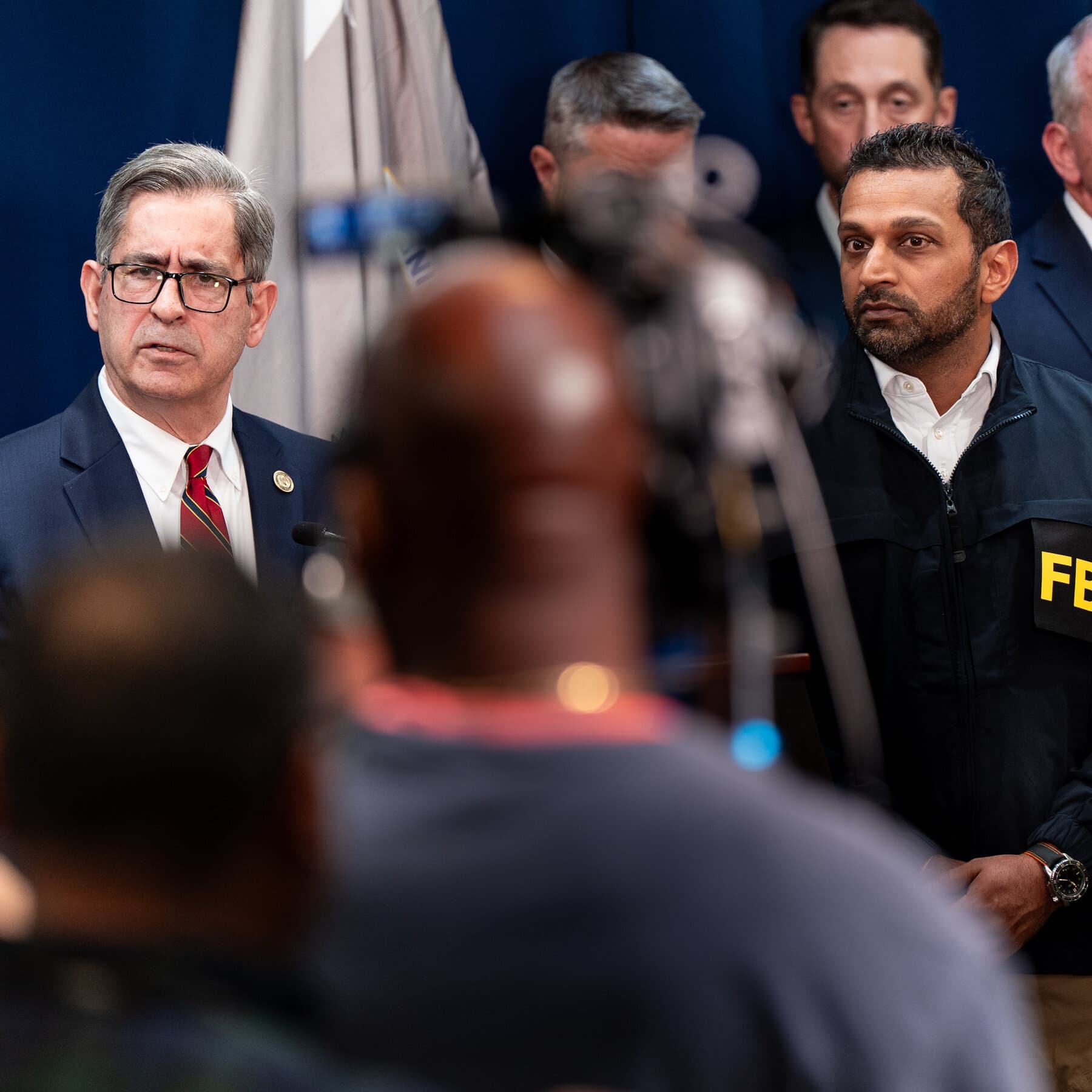 A minimalist, dark-themed digital art piece showing a professional sports arena where the grass is made of green-tinted hundred-dollar bills and the scoreboard displays fluctuating betting odds instead of points. In the center, a faceless athlete in a jersey stands like a chess piece, with glowing neon wires connected to a smartphone held by a giant, translucent hand in a suit.