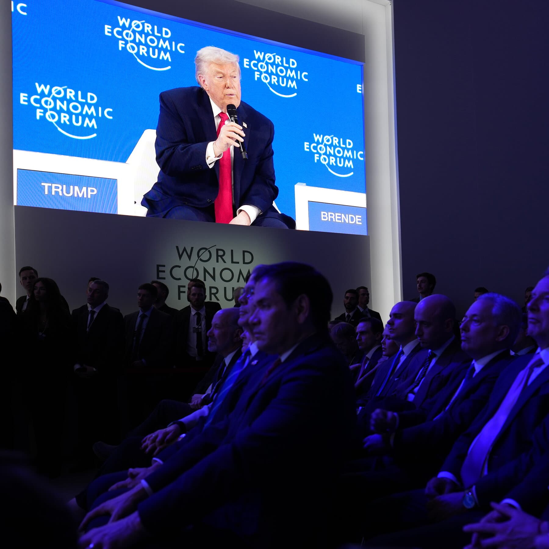 A satirical political cartoon style image featuring a giant, orange-tinted figure in a long suit and tie planting a tiny flag labeled 'MINE' on a massive, melting ice cube. In the background, small figures in European suits sip coffee and look confused. The setting is snowy mountains, cold blue and white tones.