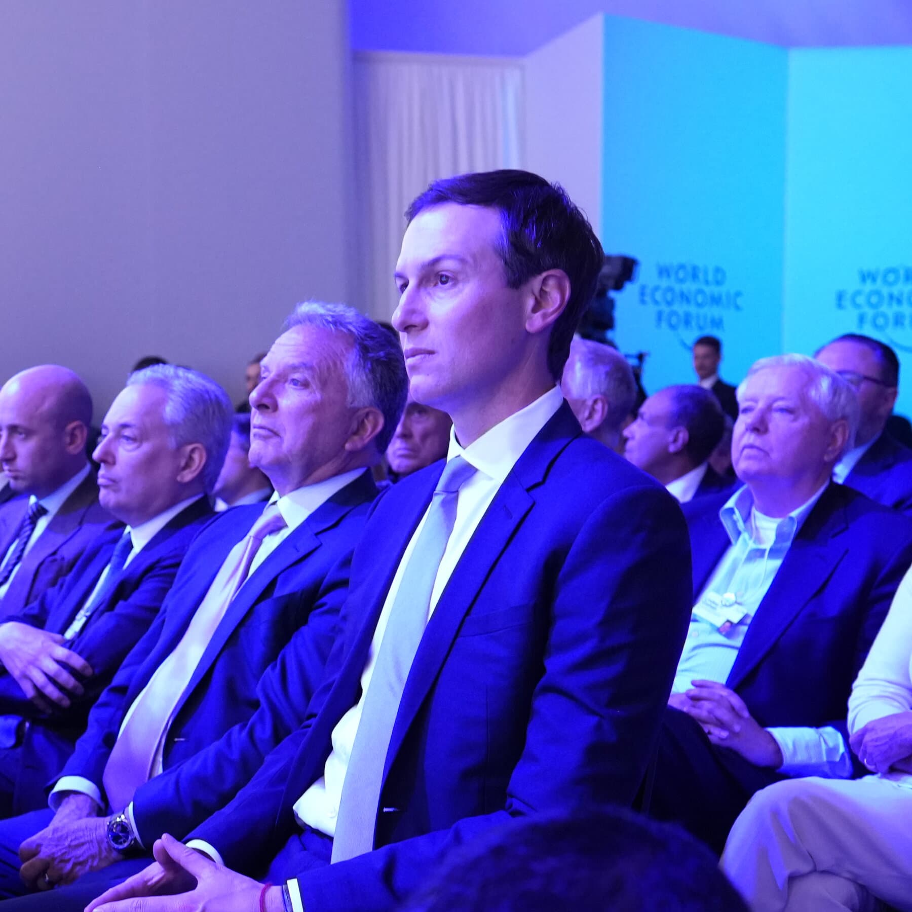 A satirical, high-contrast illustration in the style of a gritty political cartoon. In the center, a long, ornate Kremlin table stretches into the distance. At the far end sits a shadowy, menacing figure resembling Putin. In the foreground, sitting on folding chairs, are two slick, generic American businessmen in expensive suits; one holds a roll of blueprints labeled 'Condos,' the other looks like a mannequin. The room is dimly lit, with the atmosphere of a cold, transactional business deal. No text.