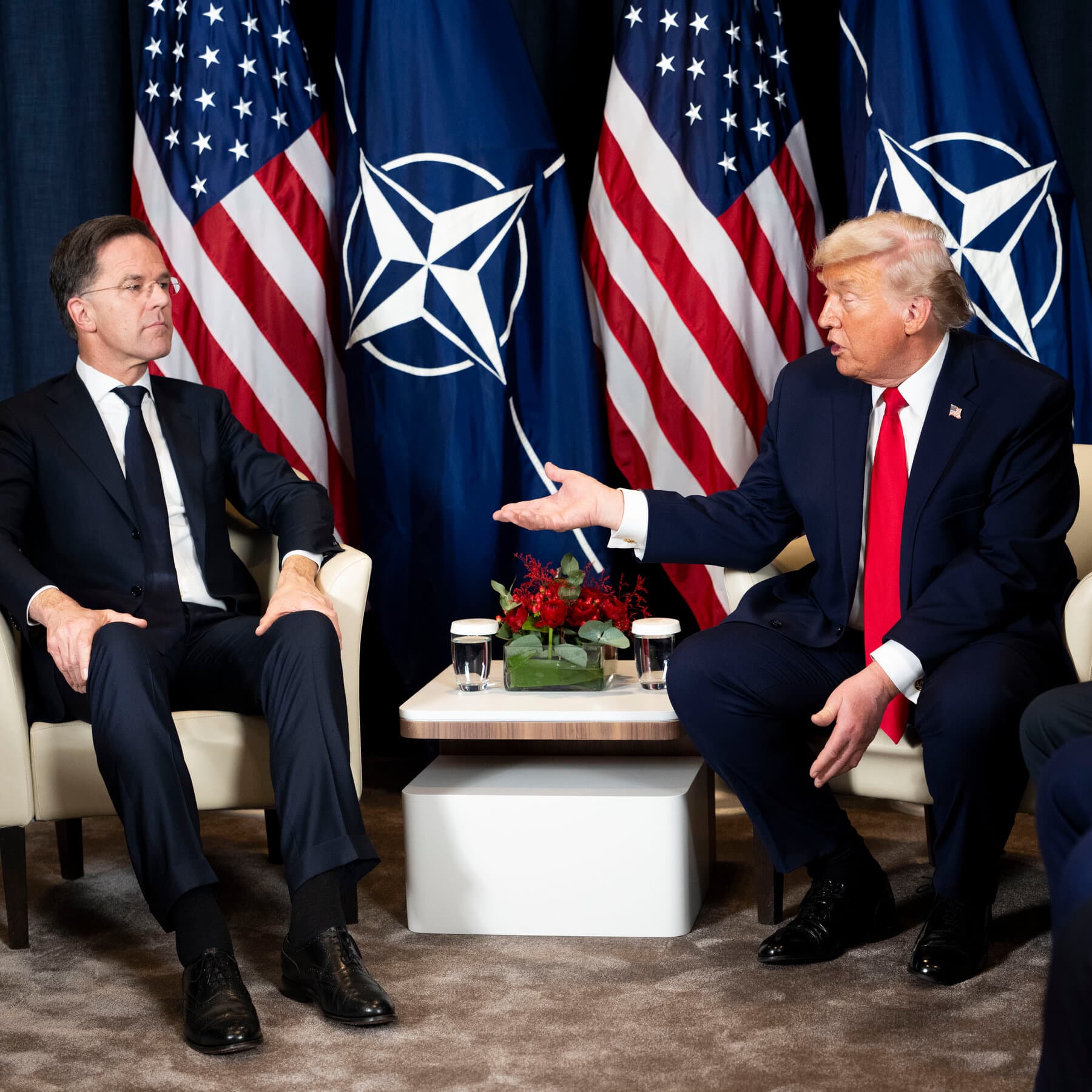 A satirical political cartoon showing Donald Trump as a 19th-century colonial explorer, wearing a pith helmet with a 'MAGA' logo, standing on a giant melting ice cube shaped like Greenland. Behind him, a group of nervous NATO officials in suits are holding a blueprint titled 'The Cyprus Loophole,' while a British redcoat from the 1960s points towards a tiny, fenced-off patch of sand on a Mediterranean beach. The style is detailed, cynical, and acid-toned, with cold Arctic blues clashing with Mediterranean teals.