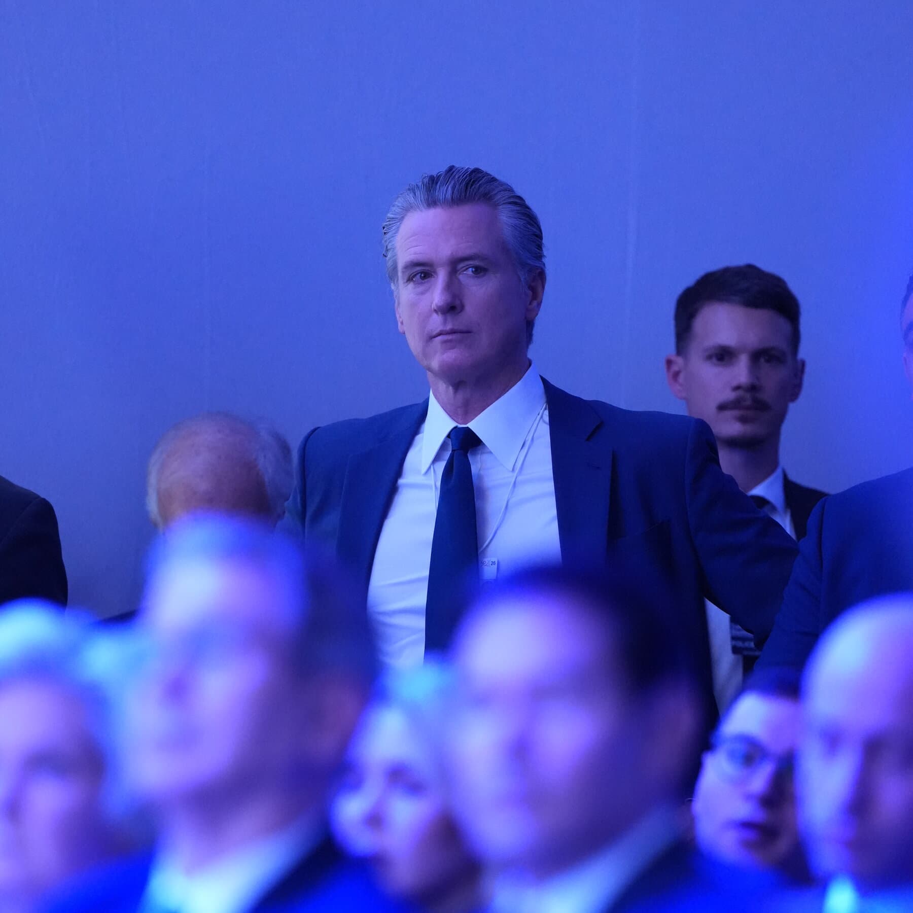 A hyper-realistic, satirical illustration of Gavin Newsom standing alone in heavy snow outside a warm, glowing Swiss chalet. He looks dejected and is holding a VIP laminate badge that says 'ACCESS DENIED'. Inside the window, silhouettes of men in suits are laughing. The atmosphere is cold, blue, and cynical.