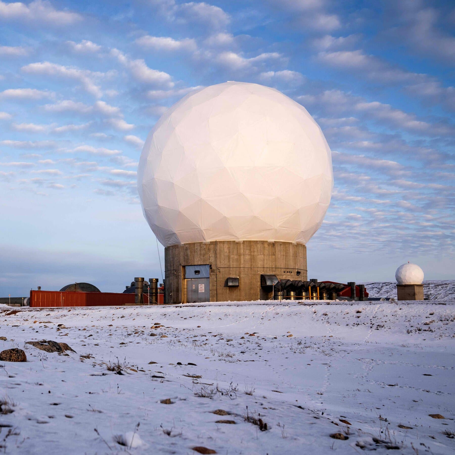 A gritty, wide-angle shot of a lone, rusty American military outpost in the middle of a vast, flat Greenland ice sheet, with a golden 'Trump' sign being hoisted by a crane in the background, overcast gray sky.