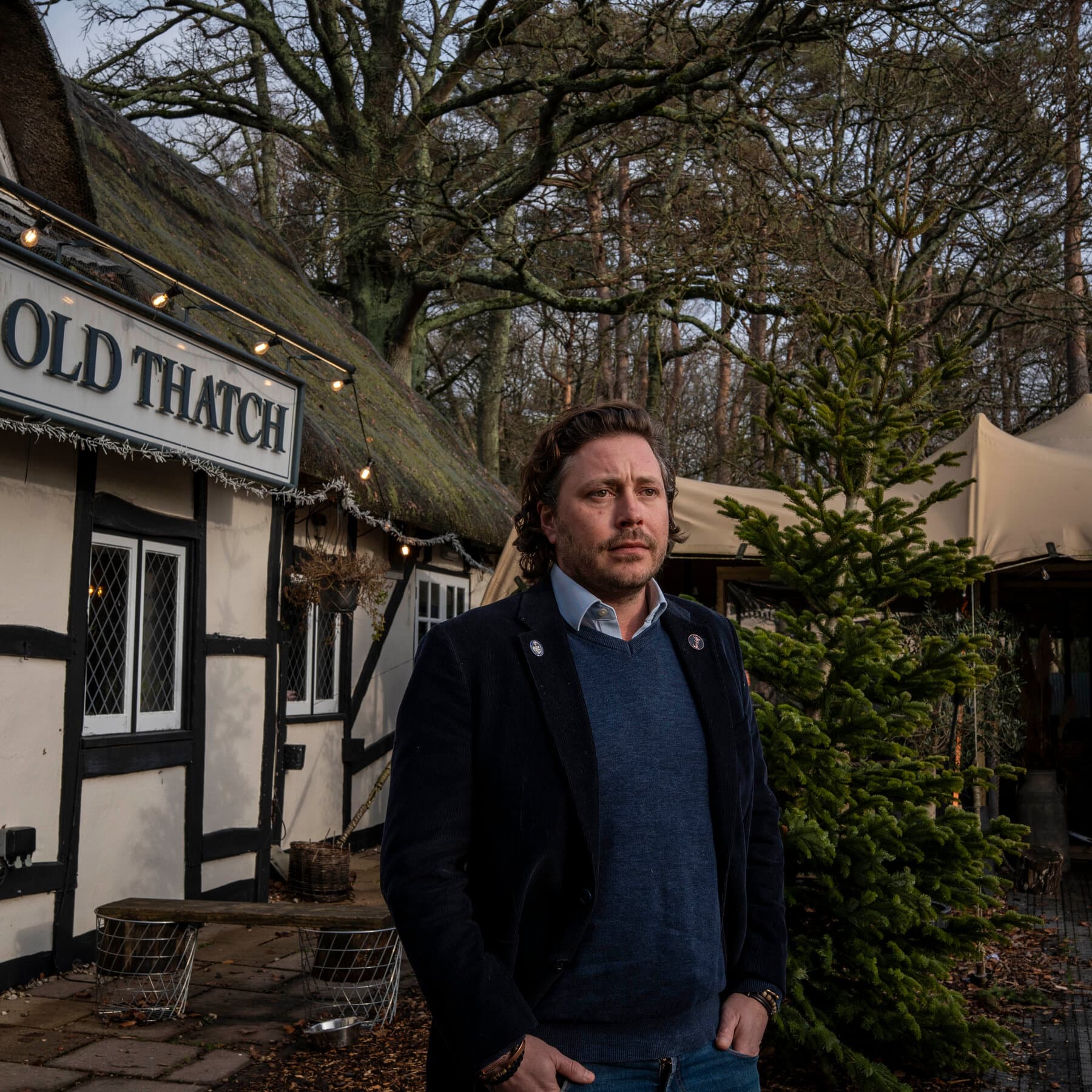 A dreary, rain-slicked British street scene focusing on the exterior of a traditional pub. A chalkboard sign stands on the sidewalk with handwritten text reading 'NO POLITICIANS ALLOWED.' Through the window, the warm glow of the pub contrasts with the grey outside. A man in a sharp business suit with a rosette pinned to his chest stands outside looking longingly and sadly through the glass, barred from entry. The style should be slightly caricatured and moody.