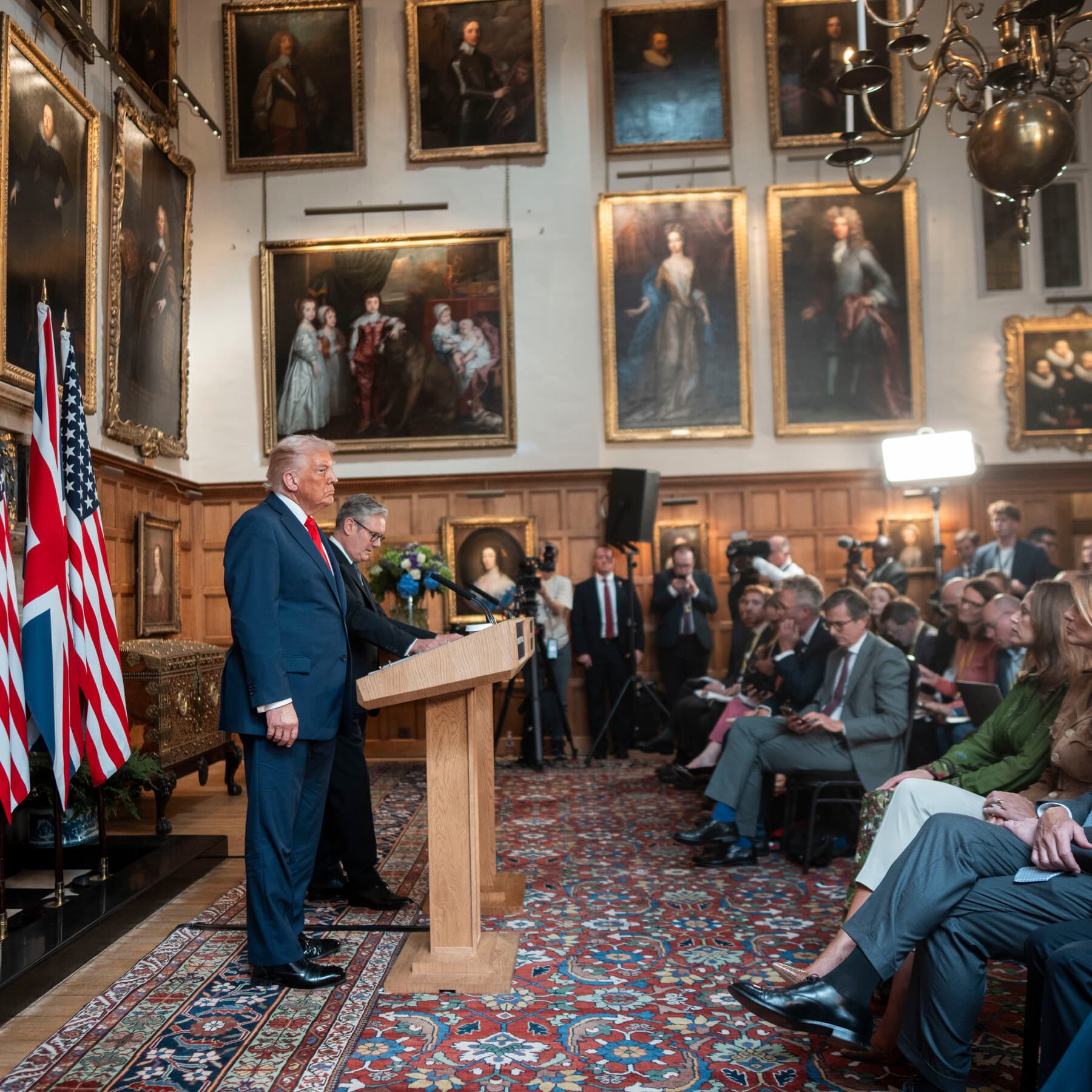A satirical political cartoon style image. A tiny, grey, exhausted-looking Keir Starmer holding a small umbrella, standing on a small map of Britain. He is being blown backward by a massive, orange-tinted hurricane wind coming from the left side of the frame. The wind has the faint shape of a shouting face. The background is a bleak, rainy London sky. The mood is cynical and overwhelming.