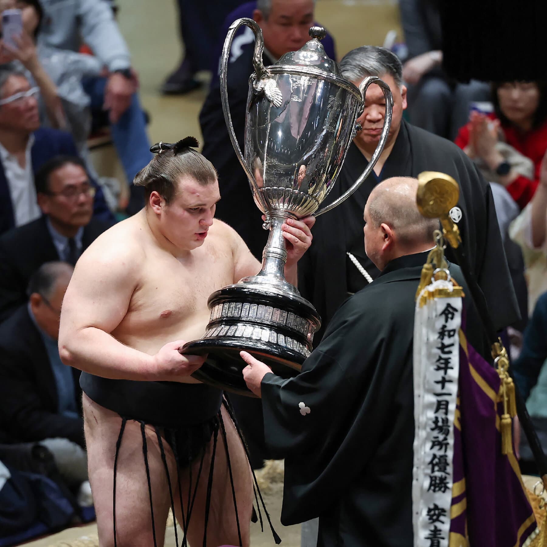 A satirical, high-contrast political cartoon style illustration showing a modern Japanese businesswoman in a sharp suit standing just outside a wrestling ring made of sand. Inside the ring, two giant sumo wrestlers look at her with confusion. The woman is holding a heavy trophy but looks hesitant to step over the line of salt. The background is a mix of modern Tokyo skyscrapers and ancient temple roofs, symbolizing the clash of eras.