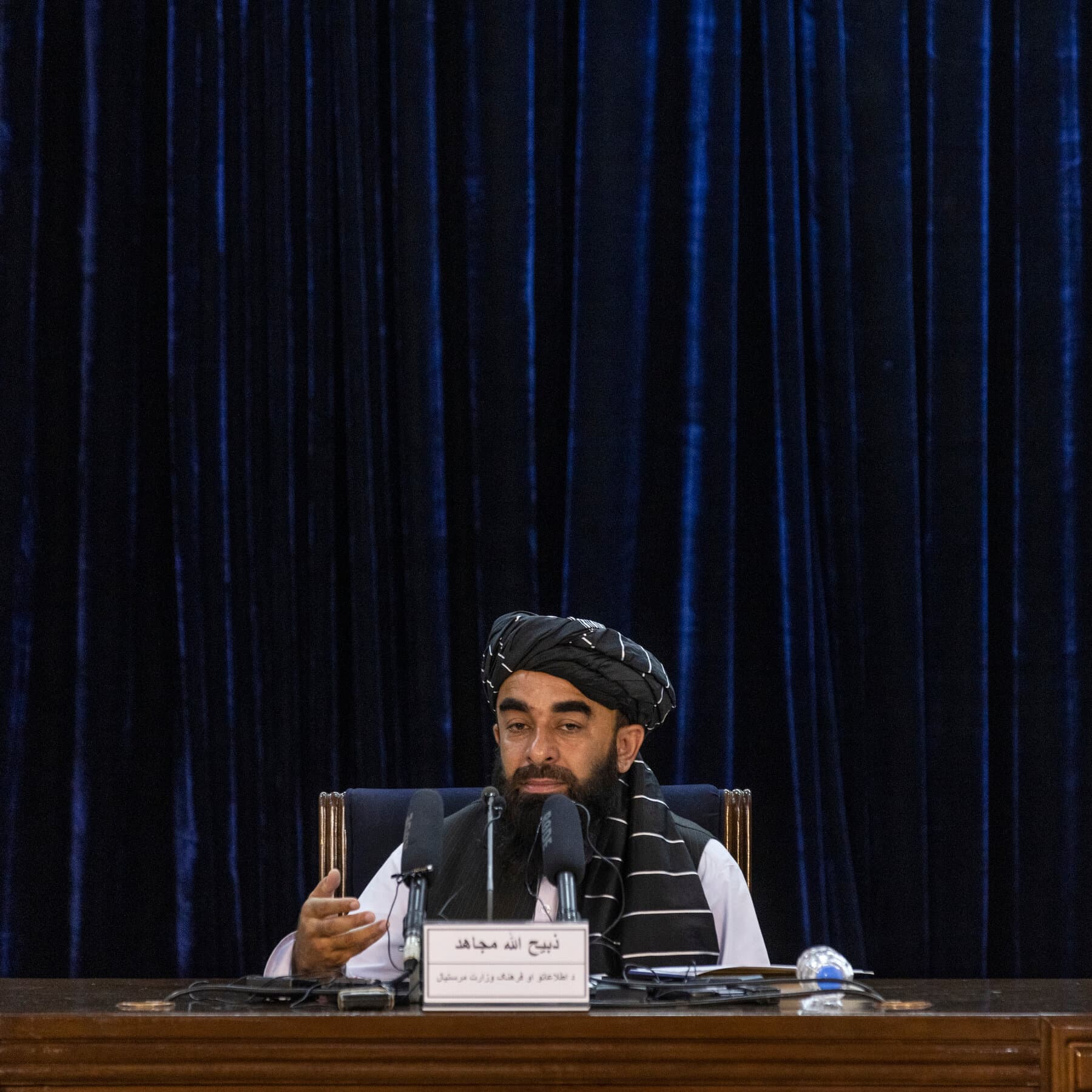 A minimalist, cynical editorial illustration showing a poker table in a dark room. On one side, a rough, turban-wearing figure holds two playing cards with human silhouettes. On the other side, a figure in a business suit points angrily at an empty spot on the table where a third card should be. The atmosphere is smoky and grim. Muted colors, dark blues and greys.