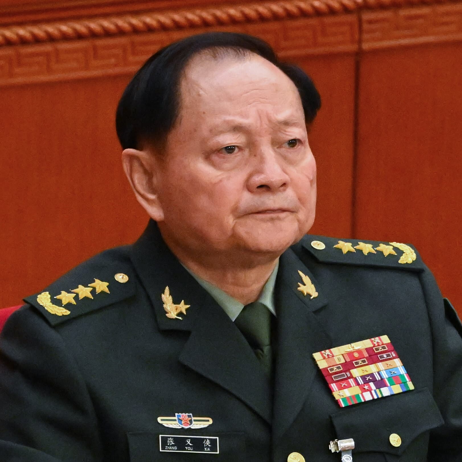 A minimalist, satirical illustration in the style of a political cartoon. A large, ornate red chair with a golden crown on it sits alone in a dark, empty room. On the floor in front of the chair, a military general's cap lies crushed and discarded. Shadows loom on the walls, creating a sense of isolation and paranoia. High contrast, muted colors.