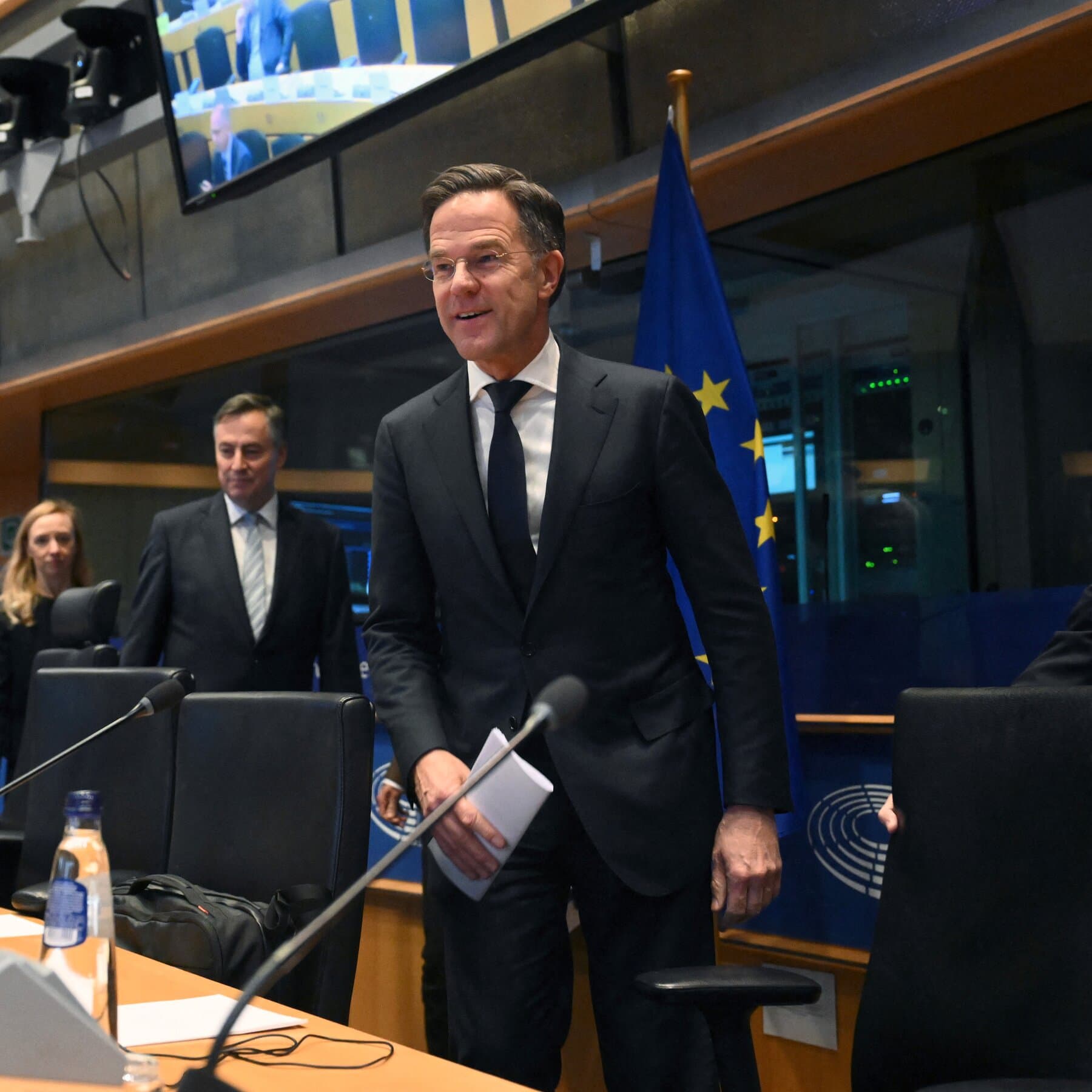 A hyper-realistic, satirical editorial illustration showing a fragile, antique European porcelain teacup sitting on a table, cracked down the middle. In the reflection of the tea, a storm is brewing. Behind the teacup, a large, shadowy American eagle looms protectively but menacingly. The lighting is dramatic and moody, emphasizing the fragility of the cup.