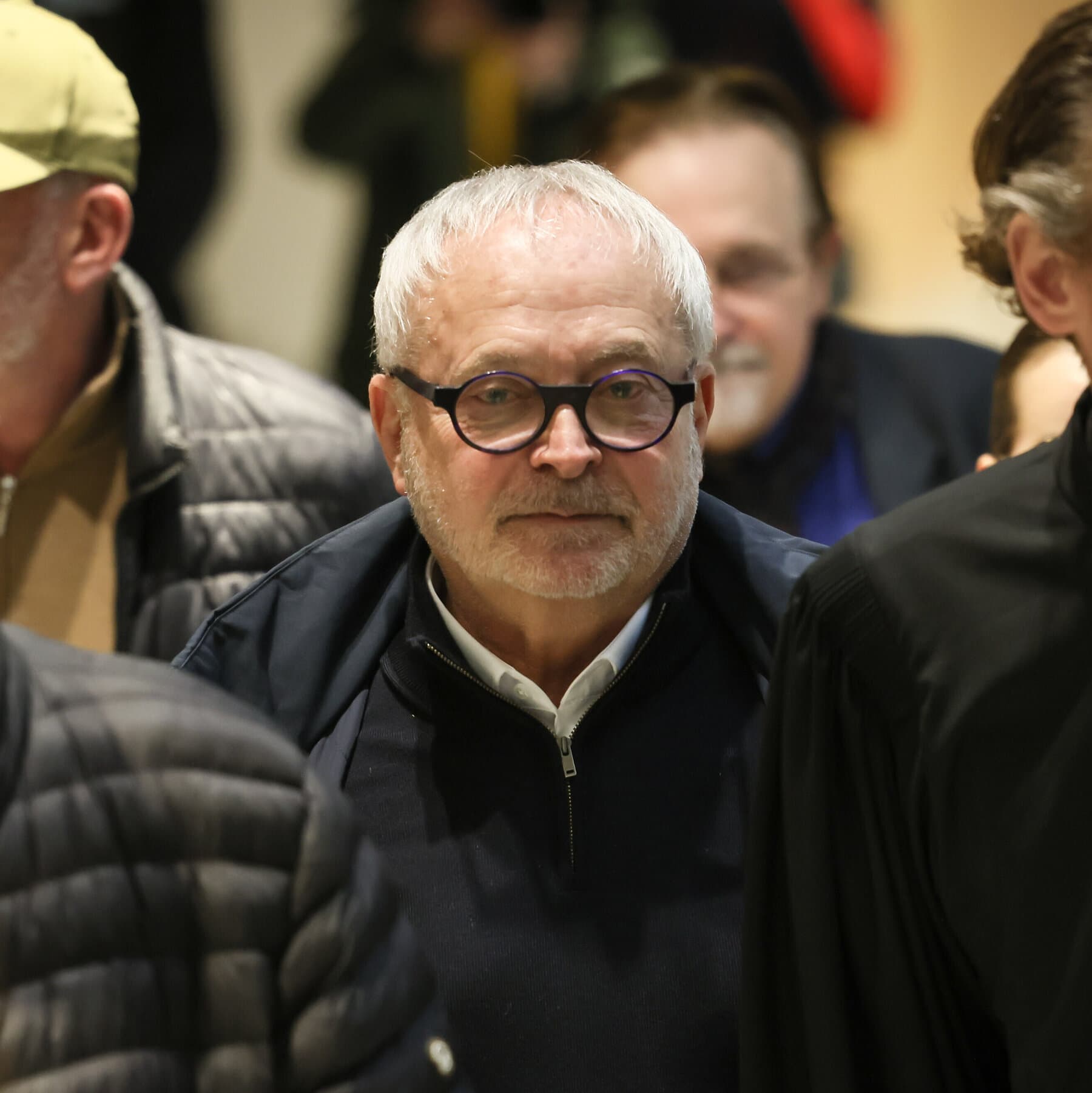 A gritty, noir-style close-up of a champagne glass on a fancy wooden desk, with a small white pill dissolving in the bubbles. In the blurry background, a man in a dark suit stands in the shadows. High contrast, cynical atmosphere.