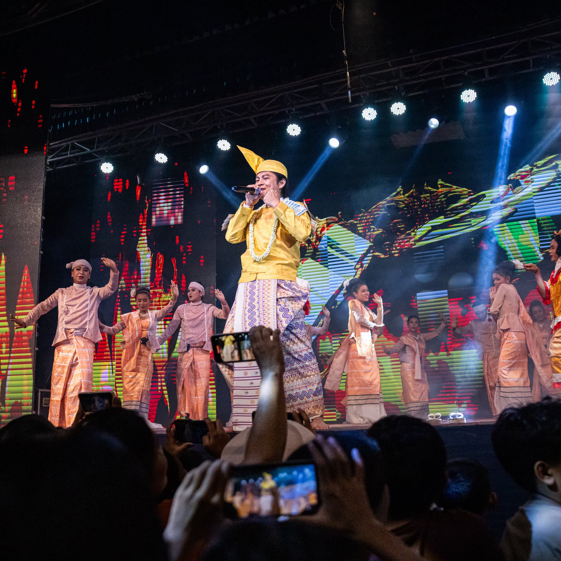 A satirical political illustration showing a bright, colorful traditional Burmese stage with dancers in elaborate costumes smiling forcefully. In the foreground, the backs of military generals in dark green uniforms are watching the show. Outside the edges of the bright stage, the background is dark, grey, and crumbling, depicting a city in ruin with smoke rising, contrasting the fake joy of the performance with the grim reality surrounding it.