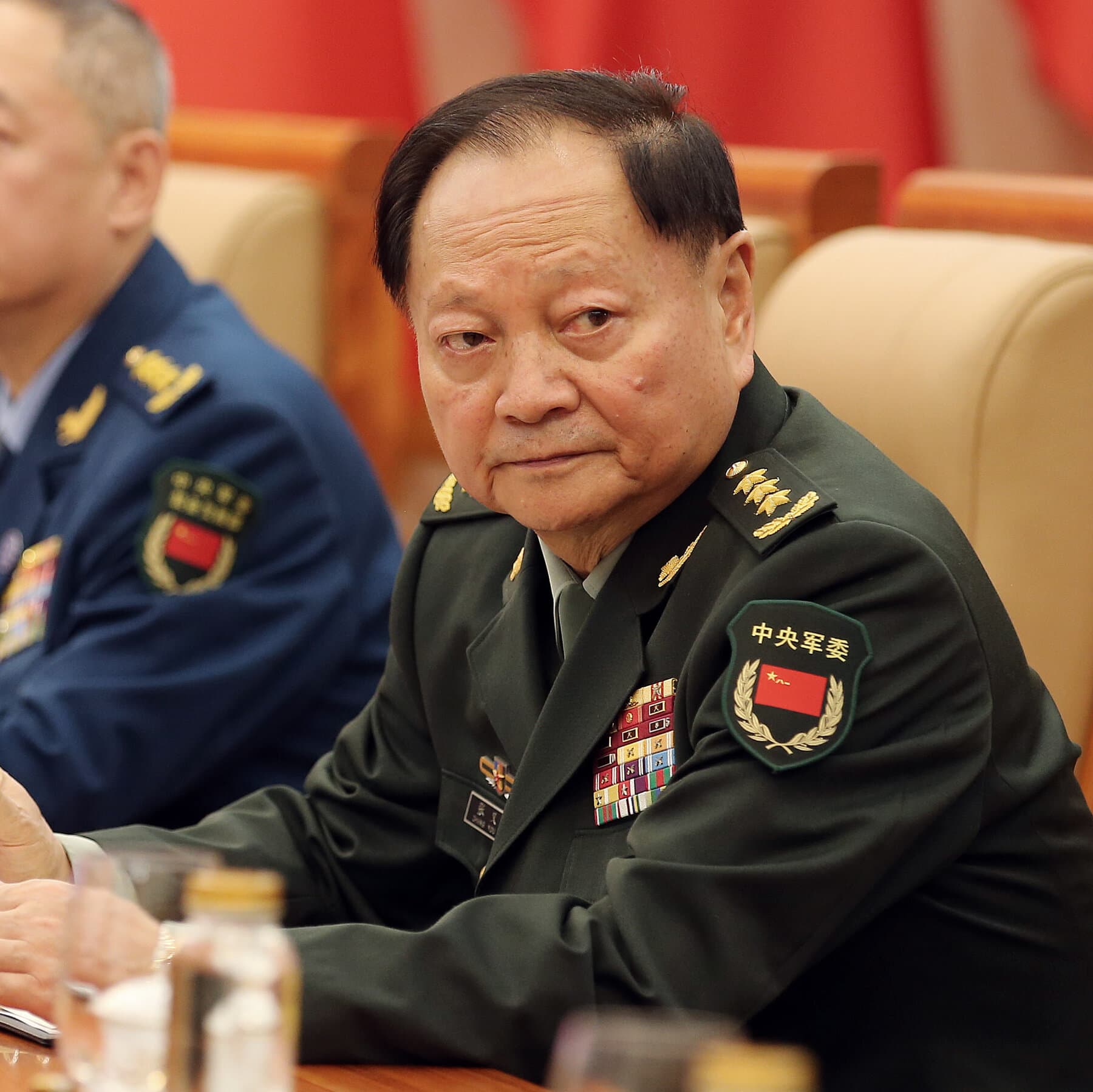 A single, empty, ornate wooden chair sitting in the center of a vast, dark room with red curtains. A military cap and a medal lie abandoned on the polished floor in a spotlight, while long shadows stretch across the scene.