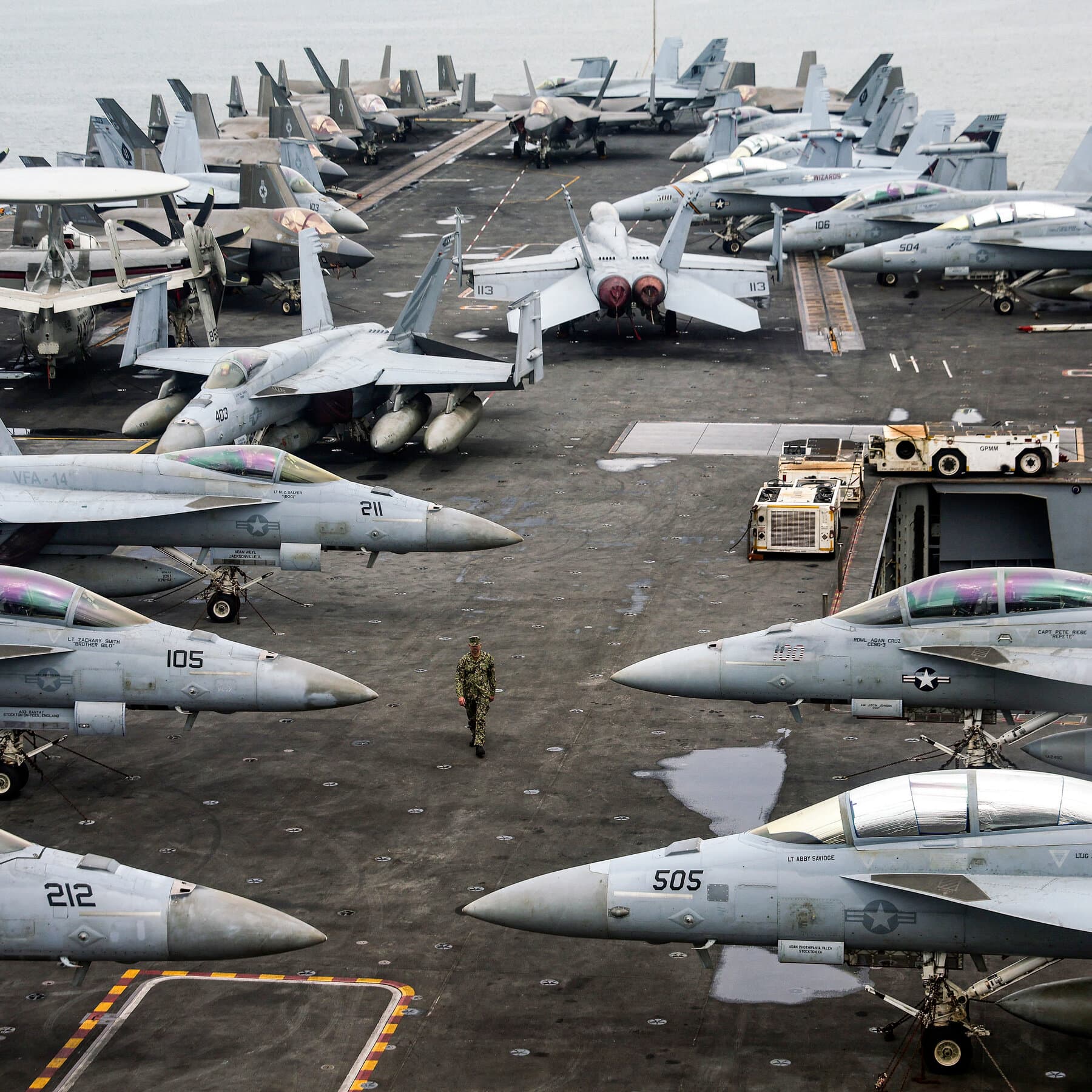 A satirical, dark political cartoon style illustration. In the center, a vast, dark ocean. On the water, a cluster of oversized, toy-like battleships bobbing clumsily, labeled 'THE ARMADA'. Above them, a giant hand emerges from the clouds holding a piece of paper labeled 'IMPOSSIBLE DEMANDS'. The sky is a gloomy grey-green. In the distance, a small cliff edge representing Iran with a 'No Trespassing' sign. The style should be ink and watercolor, heavy shadows, conveying a sense of absurd, dangerous theater.