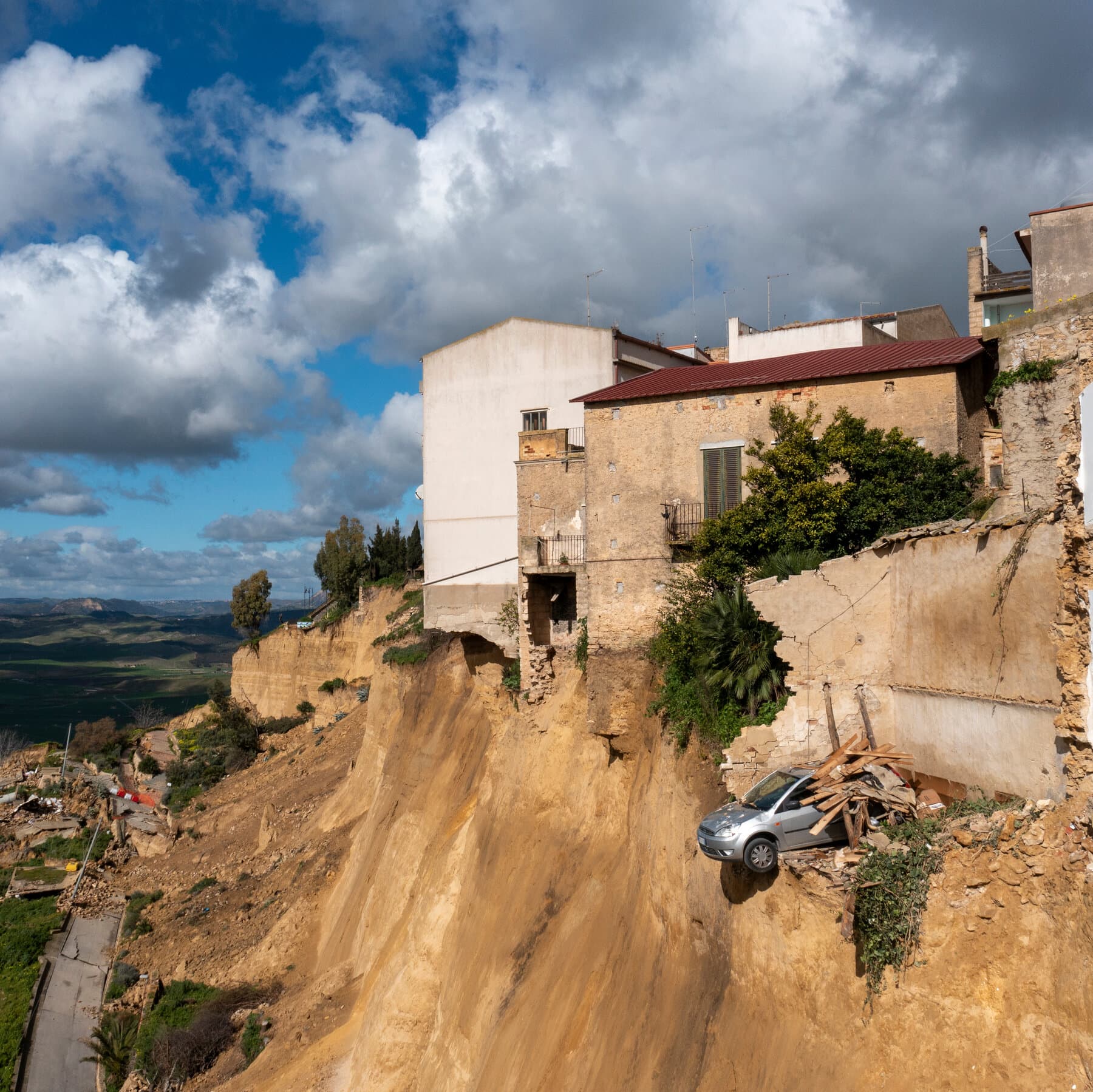 A hyper-realistic, slightly desaturated photo of a cracked Italian cobblestone street in a historic town. The street ends abruptly at a jagged cliff edge where the earth has fallen away. In the background, thick fog obscures the drop. Yellow caution tape flutters uselessly in the wind. The lighting is overcast and moody, emphasizing the texture of the broken asphalt and old stone.