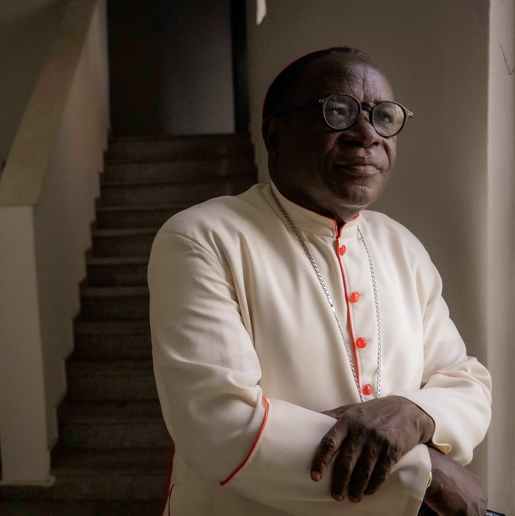 A weary Catholic bishop in simple robes standing alone on a dusty Nigerian street, looking tired and frustrated, with a blurred background of a chaotic crowd and smoke, realistic style, 8k resolution.