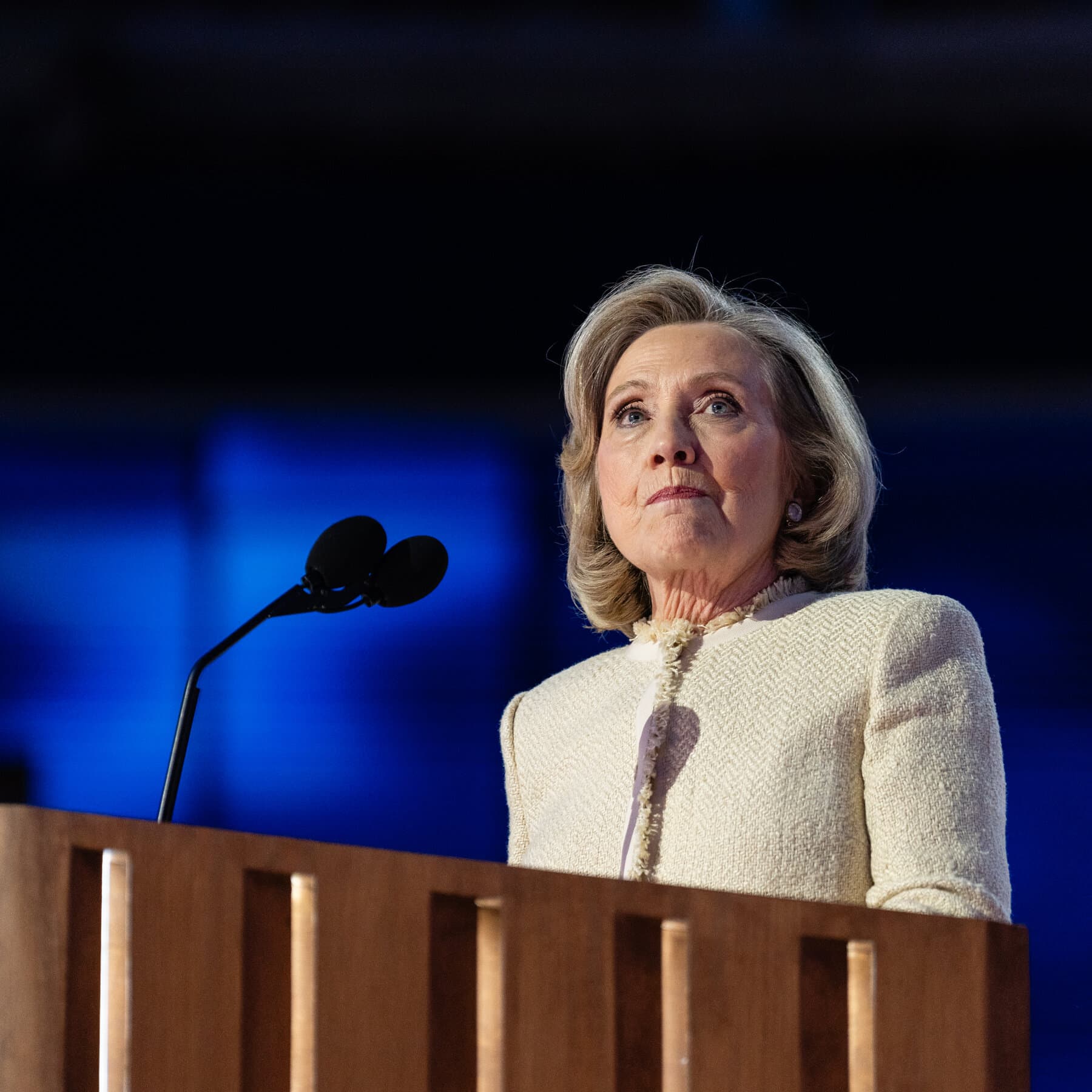 A gritty, satirical illustration in a split-screen style. On the left side, Hillary Clinton in a pantsuit speaking into a microphone with a bored expression. On the right side, Tucker Carlson with a confused expression wearing a suit and tie. In the background, instead of an American flag, there is a blurry, golden, opulent palace interior with oil wells visible through a window. The color palette should be sandy yellow, dark suit blue, and the green of money.
