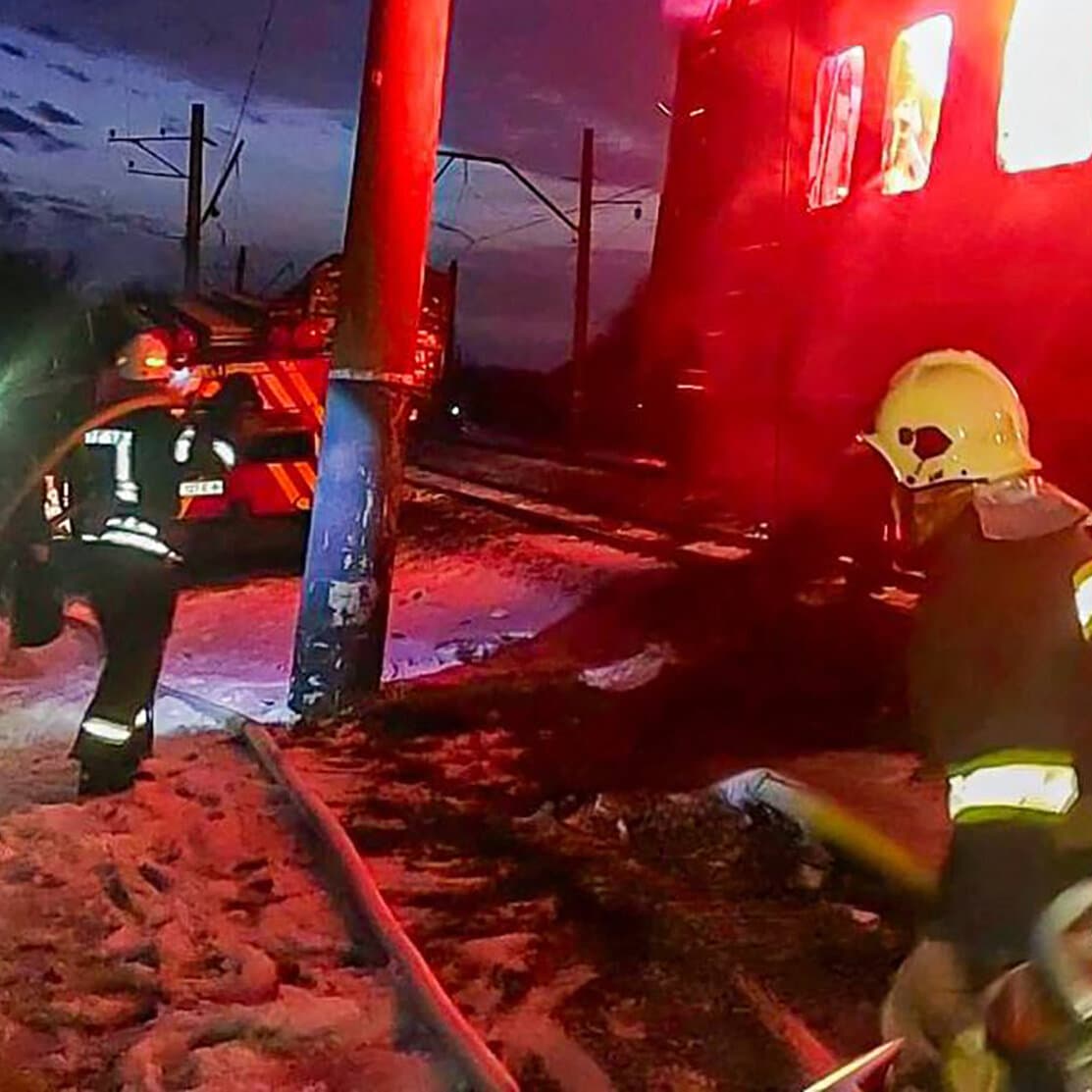 A gritty, high-contrast illustration of a train moving away into the distance on a bleak landscape. In the foreground, detached train cars are engulfed in flames and black smoke. The scene is somber and industrial.
