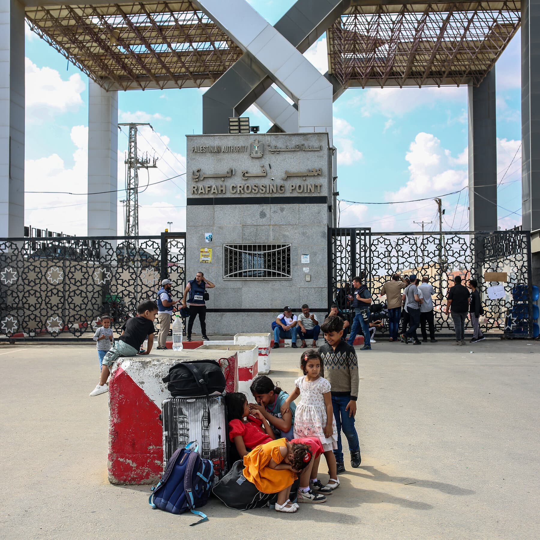 A rusty, heavy metal gate in the middle of a dusty desert. The gate is slightly open. On the left side, a man in a dark business suit stands with his arms crossed, looking angry. On the right side, another man in a dark business suit stands with his back turned, looking at a watch. In the background, a long line of white trucks waits in the heat waves. The sky is bleak and gray. High contrast, gritty, realistic style.