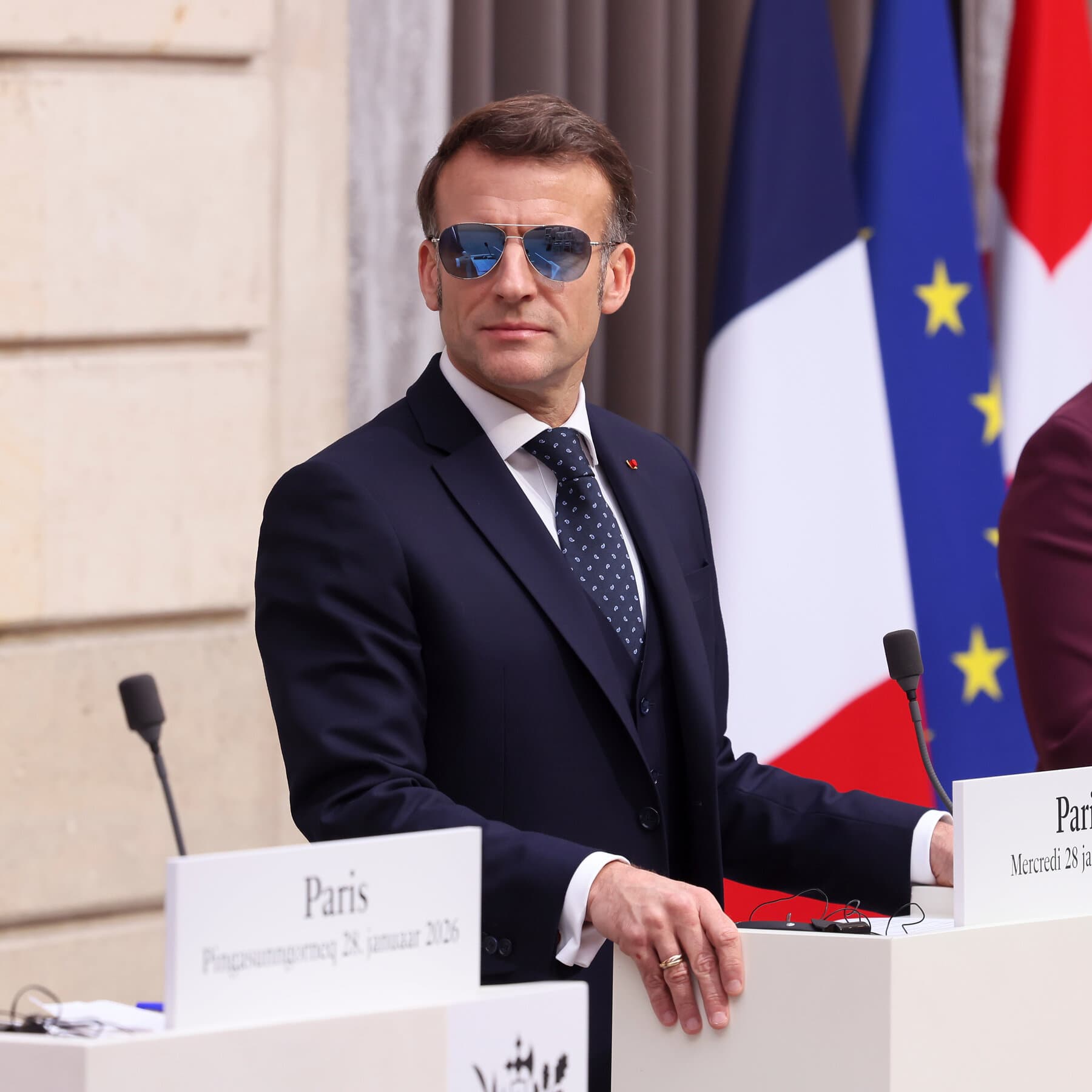 A caricature of Emmanuel Macron standing on a tiny soapbox wearing a Napoleon hat, looking smug, while a giant orange cartoon hand tries to grab a map of Greenland in the background. The style is gritty, cynical political cartoon ink drawing.