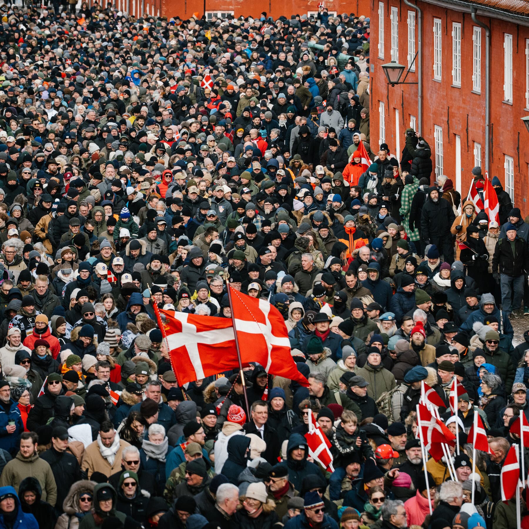 A moody, overcast street scene in Copenhagen. A group of somber veterans in civilian clothes mixed with military berets walking in a protest march. The atmosphere is grey, cold, and serious. No bright colors. Focus on the expressions of weary resignation and cynical anger on their faces. Urban European architecture in the background, slightly out of focus.