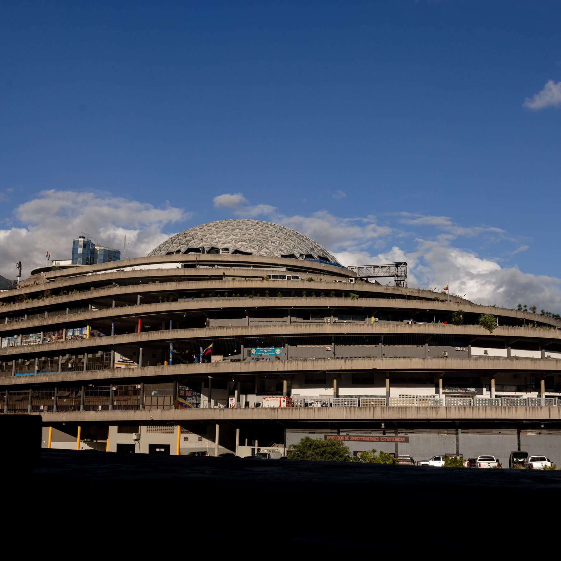 A hyper-realistic, slightly desaturated photo of a Caracas street at dawn. Faded political posters are peeling off a concrete wall. In the foreground, an empty chair sits on the sidewalk, casting a long shadow. In the background, a few blurred figures walk cautiously, looking at their phones. The atmosphere is tense and quiet.