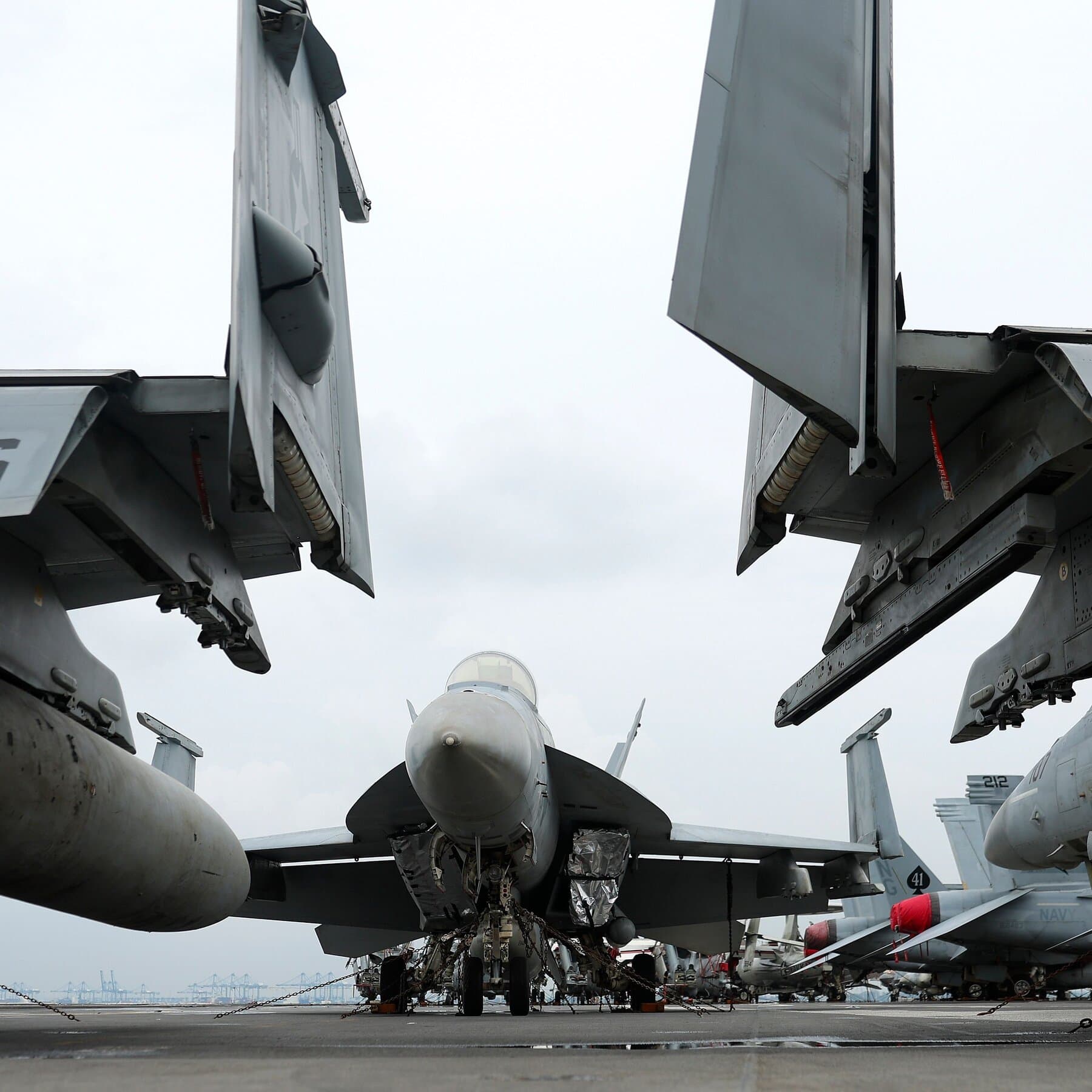 A satirical illustration showing a sleek, futuristic fighter jet firing a massive missile made of stacks of cash at a tiny, rickety toy drone buzzing around a giant aircraft carrier, set against a dramatic ocean backdrop.