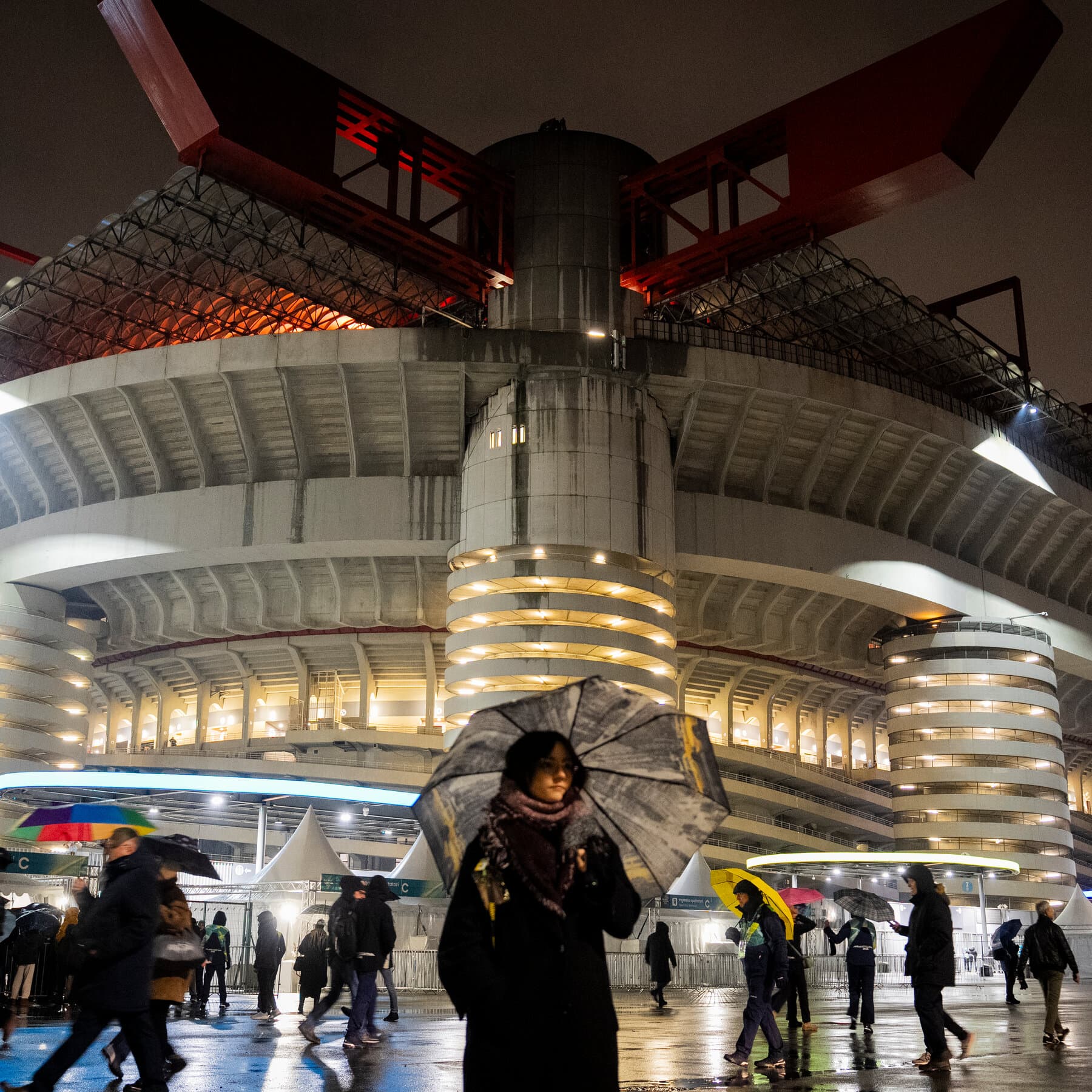 A highly contrasted, moody illustration showing a shiny, golden Olympic medal hanging from a heavy, rusted iron chain. In the background, blurred silhouettes of riot police stand in the snow. The atmosphere is cold and cynical.