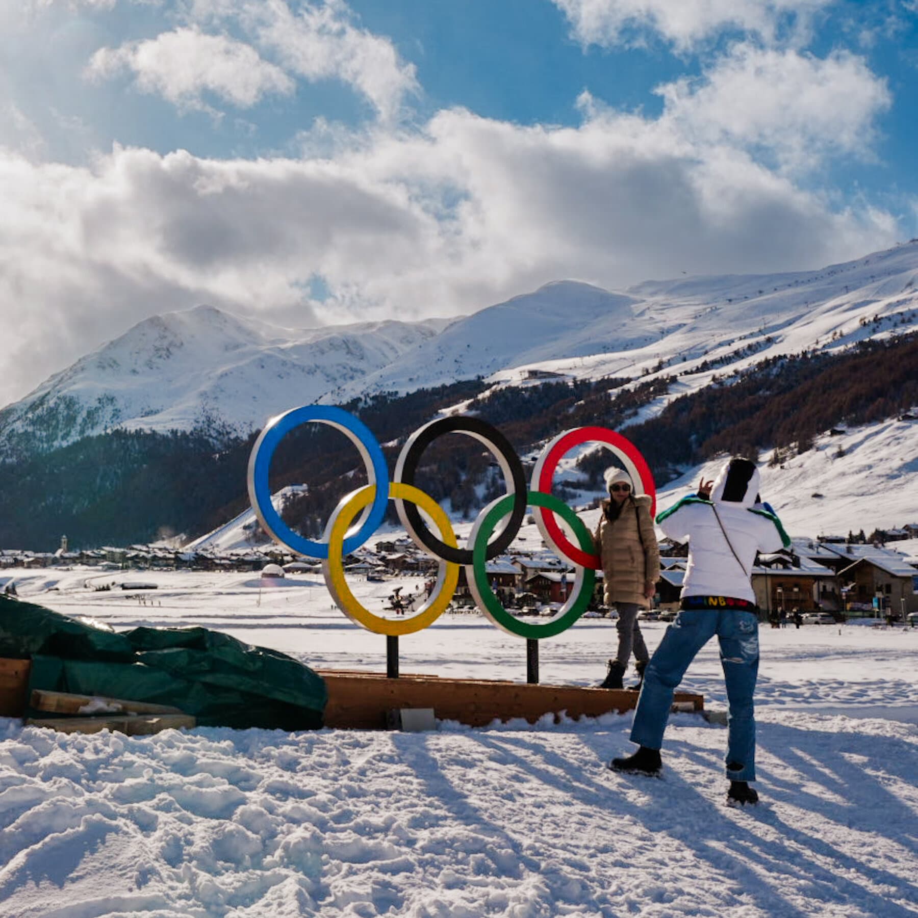 A highly stylized, cynical editorial illustration. In the foreground, a pristine, glowing snow globe sits on a cracked, dirty table. Inside the snow globe, tiny gold figures ski and skate in a perfect winter wonderland. Outside the snow globe, the background is dark and chaotic, depicting a world in disarray with storms, crumbling buildings, and red warning lights. The contrast between the perfect fake world inside the glass and the disastrous reality outside is sharp and satirical. Digital art style, muted colors for the background, bright fake colors for the snow globe.