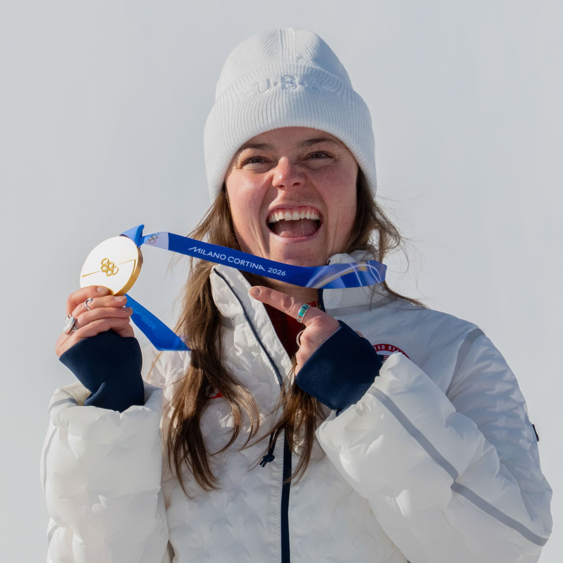 A sad, close-up photo of a gold medal lying in the dirt with a frayed, broken ribbon next to it. The background is blurry, showing the legs of an athlete standing still. The lighting is harsh and realistic.