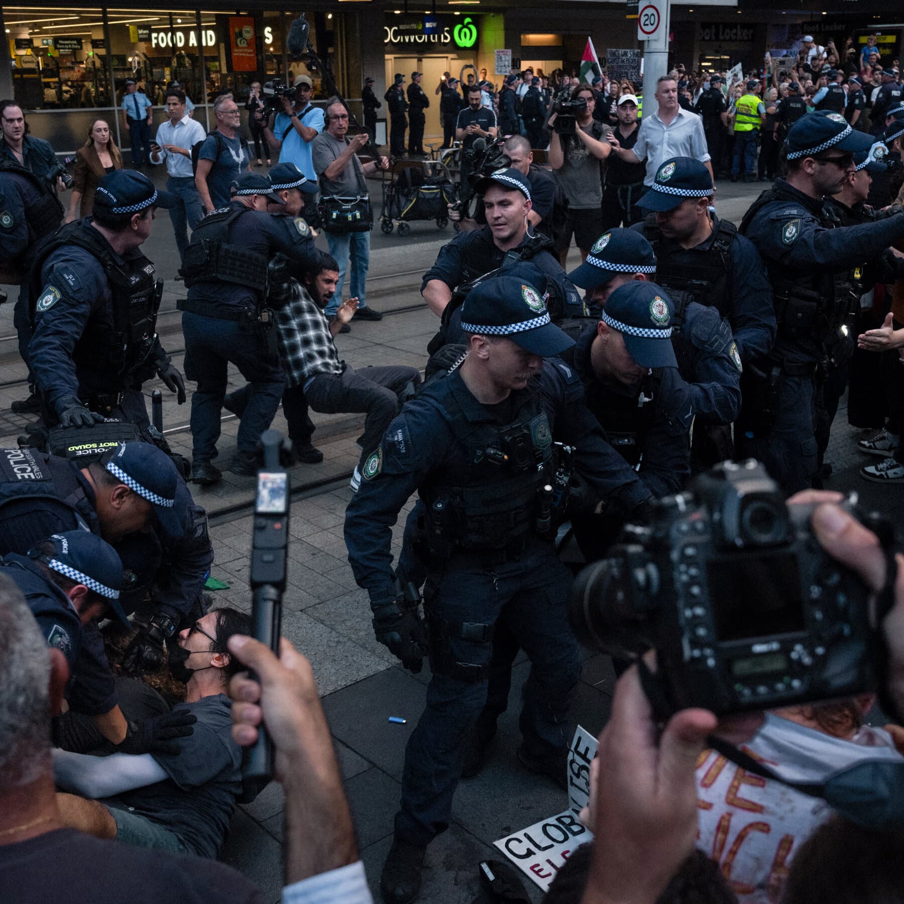 A gloomy, high-contrast illustration of a heavy metal barrier separating a blurred crowd of protesters from a line of weary police officers in Australia. In the background, a vague silhouette of a generic government building looms under a grey, stormy sky. The atmosphere is tense and cynical.