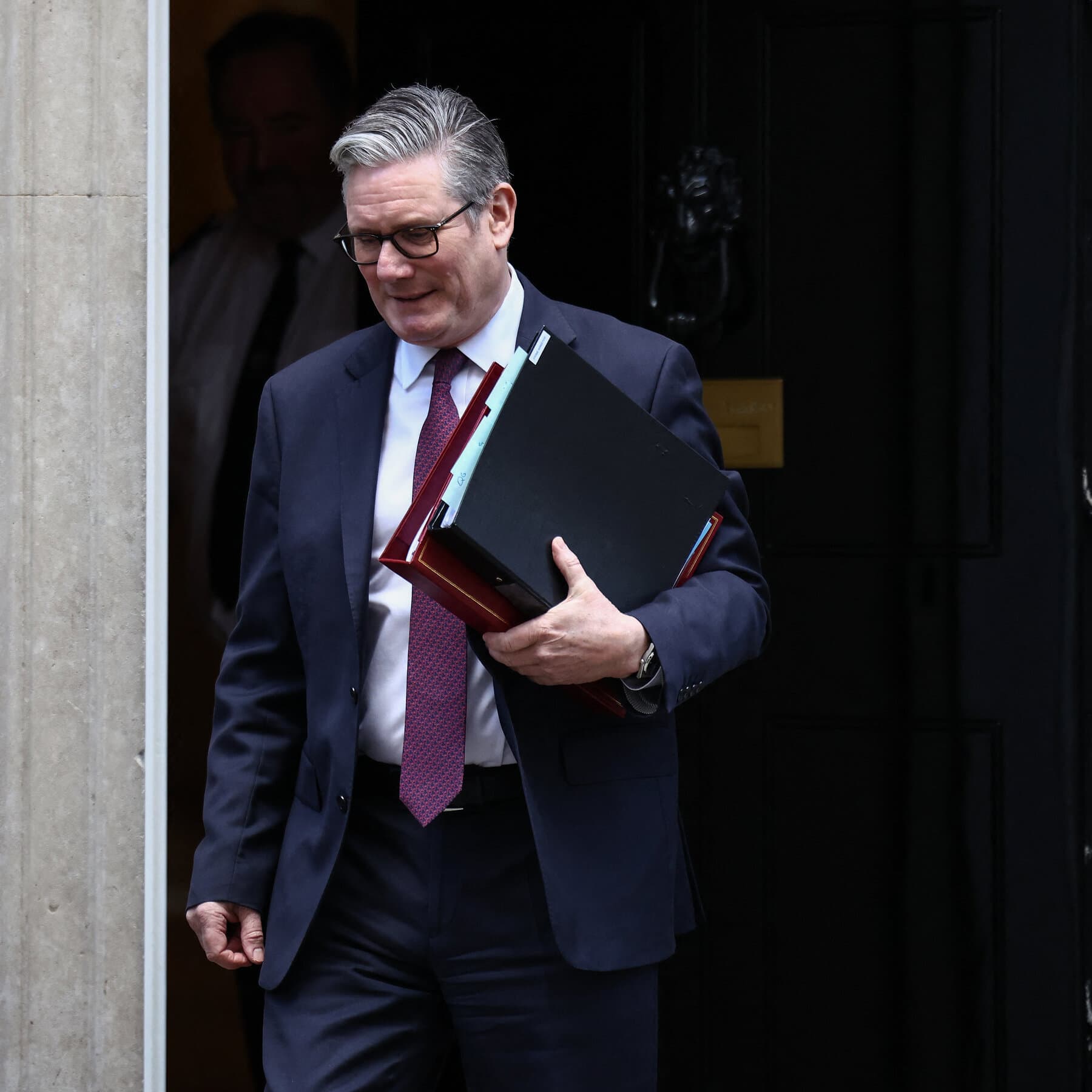 A hyper-realistic, cynical editorial illustration showing a grey, boring bureaucrat in a suit drowning in a sea of red paperwork and file folders. In the background, far away on a sunny tropical island, silhouetted figures are partying and laughing. The lighting is moody and dramatic, emphasizing the isolation of the bureaucrat.
