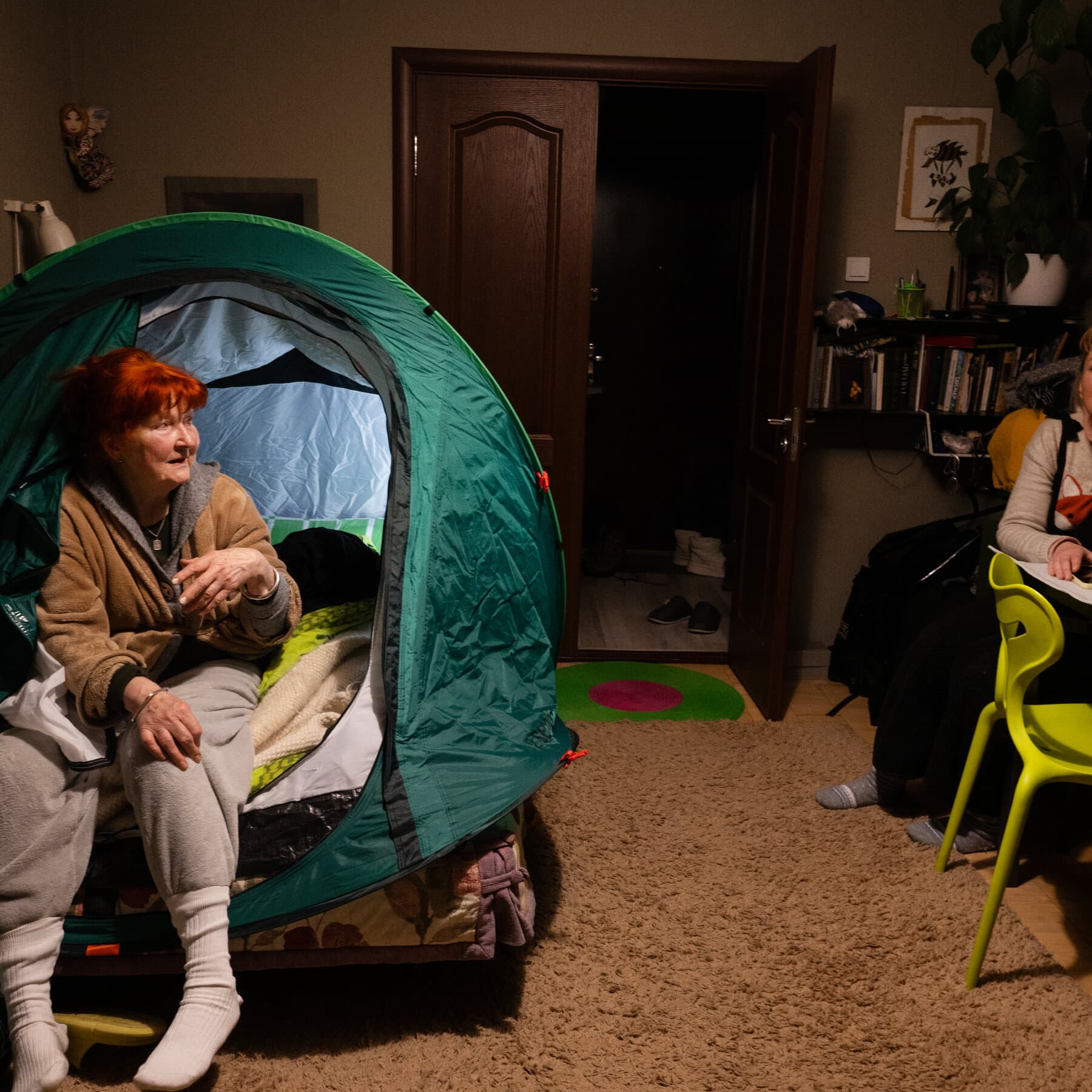 A hyper-realistic, gloomy interior of a cold apartment in Kyiv. In the center, a colorful camping tent is pitched directly on top of a double bed with blankets piled inside. The room is dimly lit by a single candle. Breath vapor is visible in the air. The atmosphere is cold, cynical, and stark.
