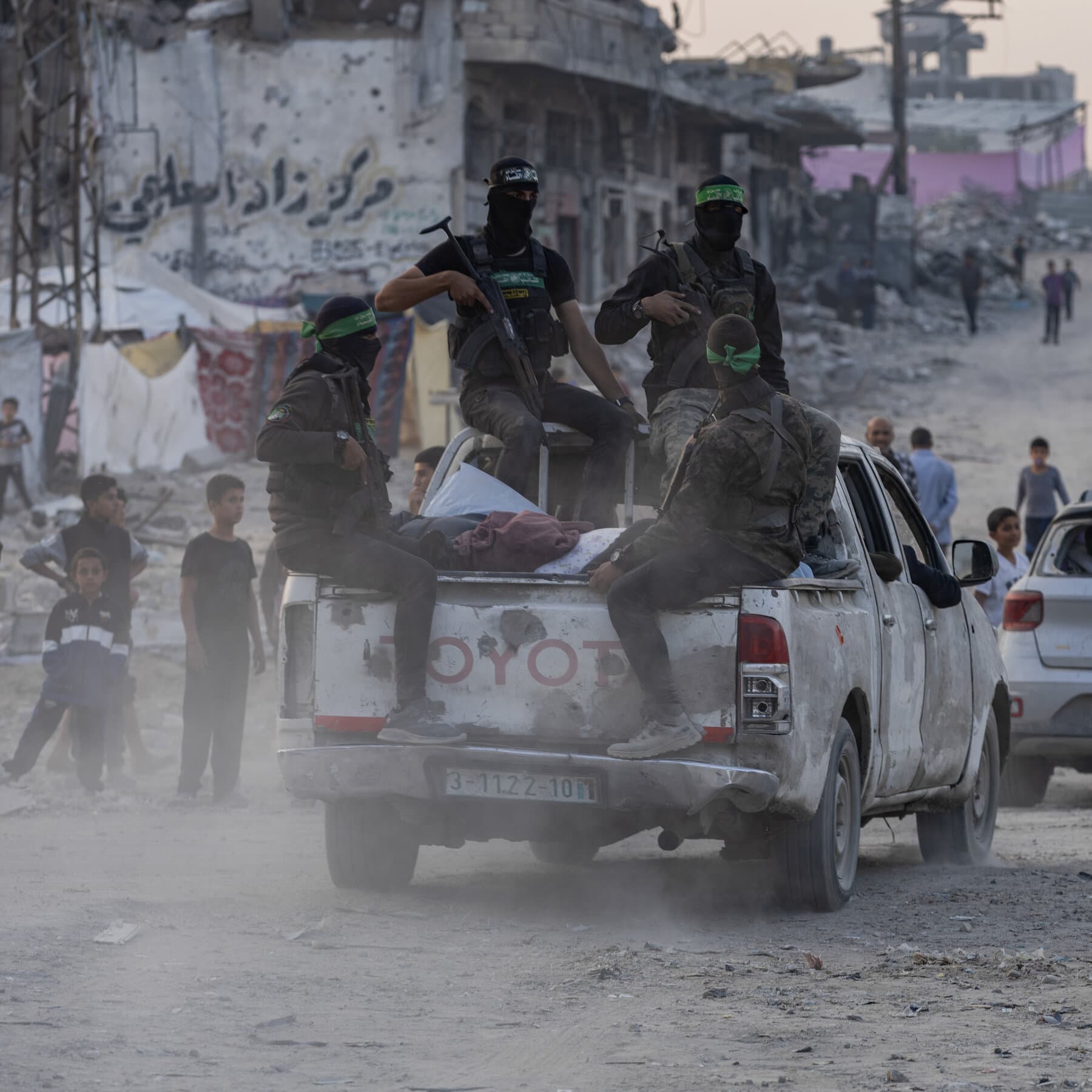 A conceptual digital illustration in a satirical editorial style. A rusty, broken cart wheel labeled 'The Plan' is rolling off an axle. The 'linchpin' is made of fragile glass or melting ice, shattering under the pressure. In the background, a blurry, chaotic desert city scene. The color palette is dusty beige, stark black, and muted red.