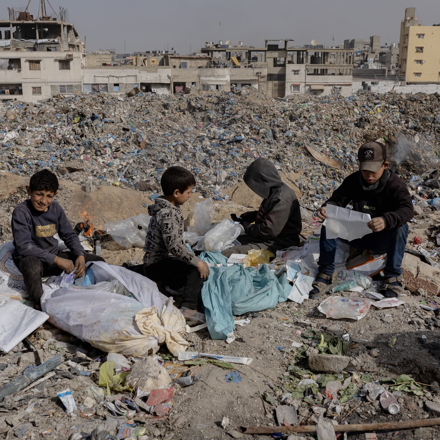 A gritty, high-contrast illustration of a golden boardroom table set in the middle of a destroyed city filled with grey rubble. On the table, stacks of green money are burning with bright orange flames. In the background, vague silhouettes of men in expensive suits are shaking hands, ignoring the fire and the ruin around them. No text.
