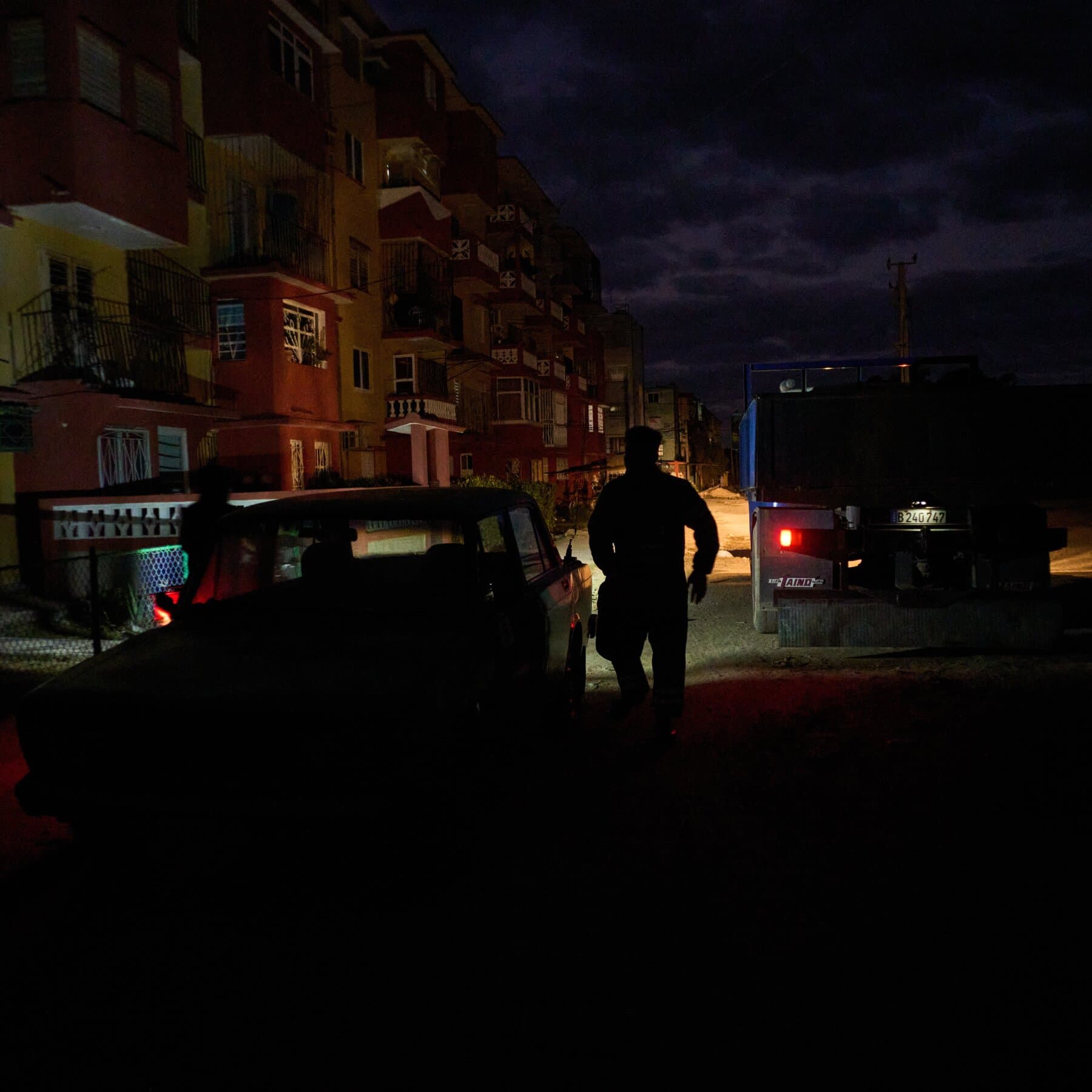 A giant, rusty oil valve with an American flag painted on it being turned off by a hand in a business suit. Below the pipe, a small, desolate island shaped like Cuba is drying up and cracking. In the background, a man in a blue UN helmet stands watching and taking notes on a clipboard, looking useless and bored. Gritty, cynical political cartoon style.