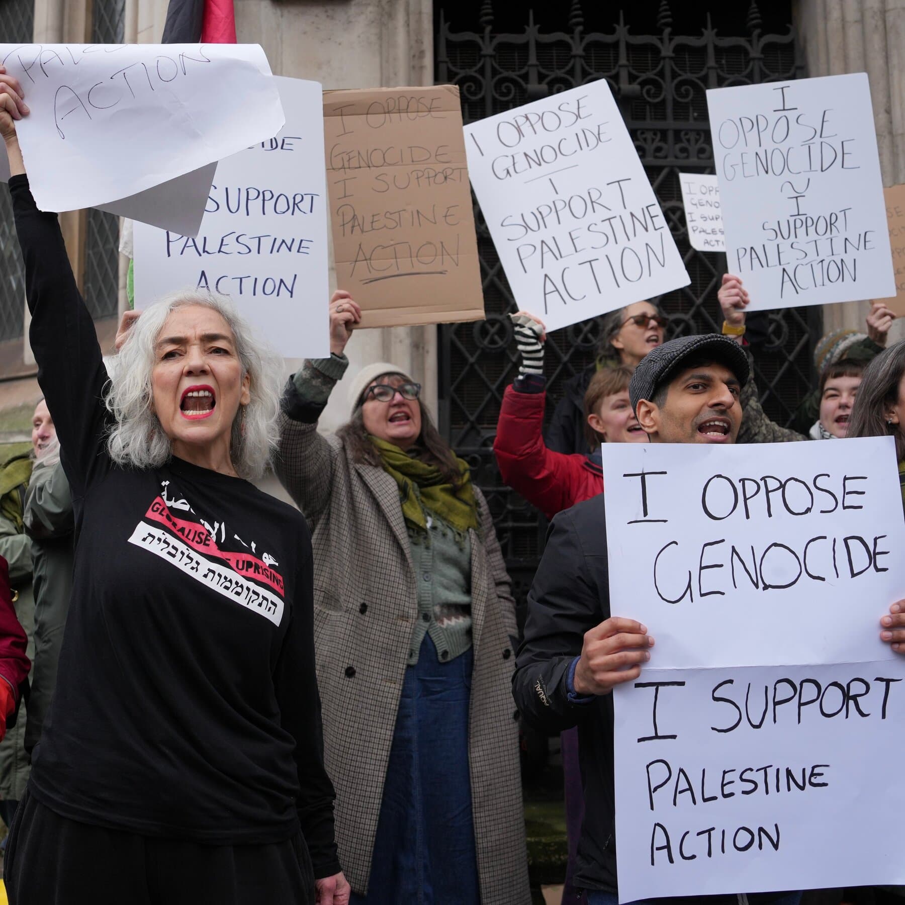 A gritty, low-angle view of a shattered glass window on a gray, industrial brick building. Through the broken glass, a blurry, desaturated British flag hangs limply in the rain. Red paint is splattered messily across the brickwork and the glass shards. The lighting is overcast and gloomy. No people are visible, only the aftermath of chaos and the cold, unfeeling architecture.