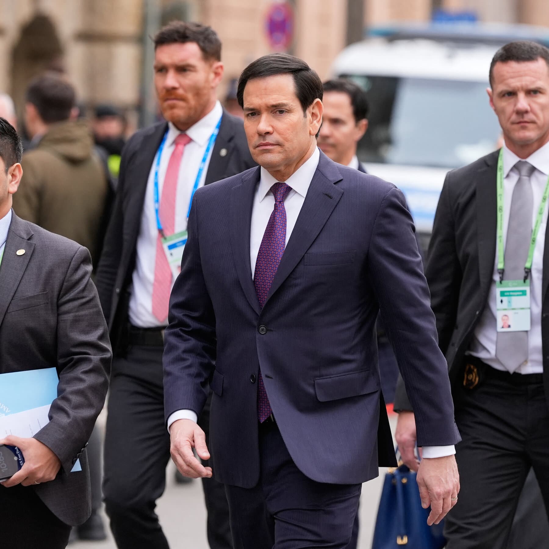 A moody, noir-style illustration of a group of nervous politicians in suits standing in the rain on a Munich street. They are looking up at a giant, chaotic storm cloud shaped like the United States. The street is wet and dark. The politicians look small and helpless. High contrast, dark colors.