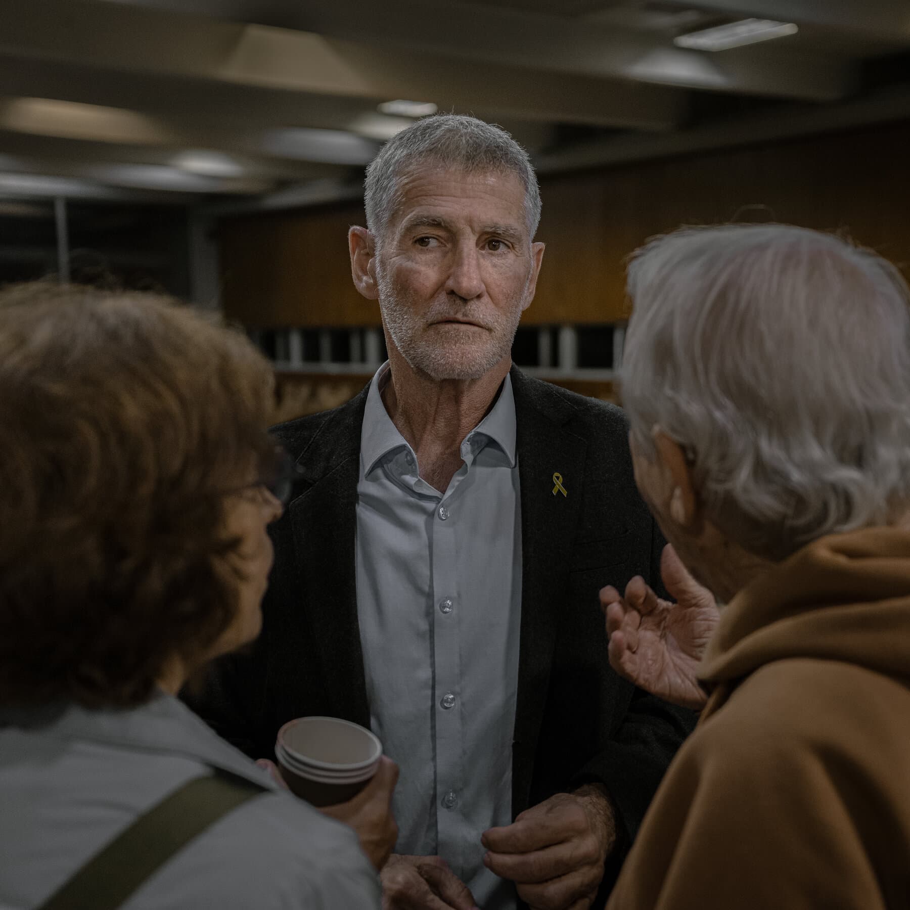 A high-contrast, gritty split image. On the left side, a pristine military general's uniform with medals shining in the light. On the right side, a dirty, torn business suit covered in mud and red paint. The background is a gray concrete wall.