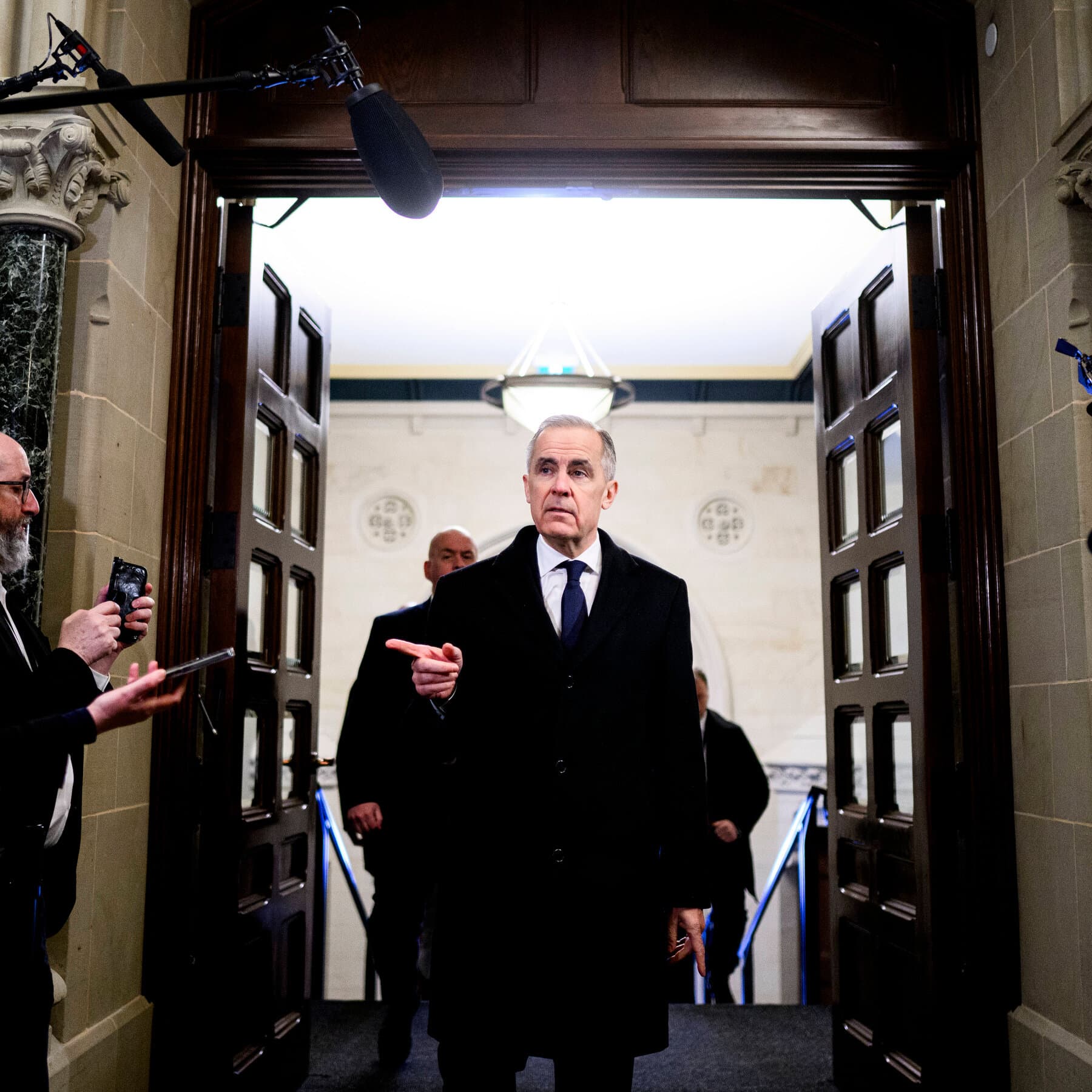 A satirical editorial illustration showing a Canadian beaver wearing a hard hat, struggling to assemble a complex, oversized tank made of wood and duct tape in the snow, while a slick, high-tech American eagle salesman in a suit watches with a skeptical expression from behind a chain-link fence. The scene is grey, industrial, and cynical.