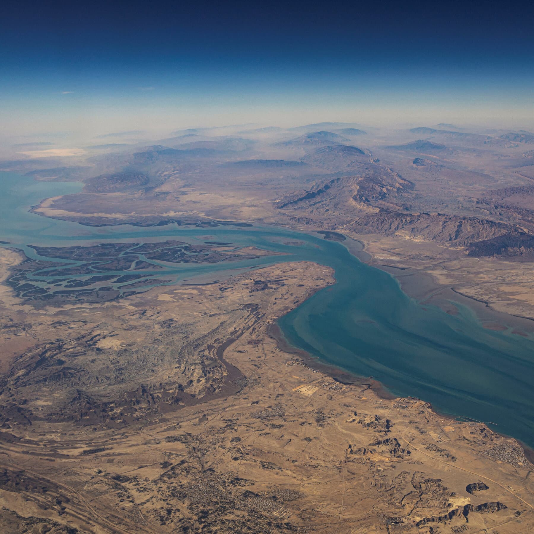 A cynical, satirical political cartoon style illustration showing a narrow body of water representing the Strait of Hormuz. On one side, a tiny table with two empty chairs and a 'Reserved for Peace Talks' sign. On the water, massive, exaggerated warships are doing donuts and splashing water everywhere, creating huge waves that threaten to tip over the table. The sky is grey and ominous. The style should be gritty and ink-washed.