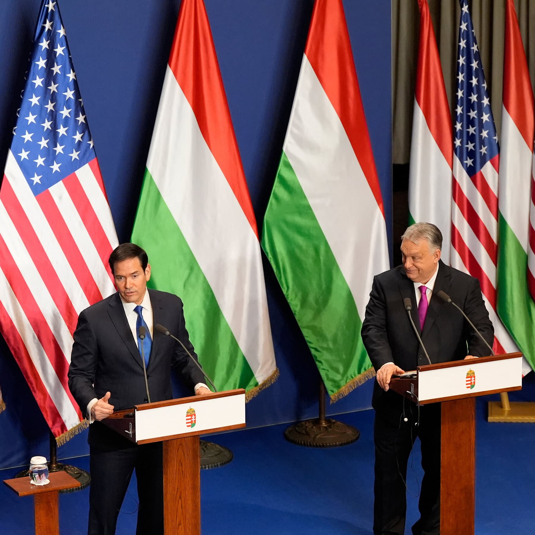 A surreal, high-contrast editorial illustration depicting a crumbling theater stage. On the stage, two empty suits shake hands; one suit is patterned with the American flag, the other with the Hungarian flag. The background is a dark, stormy sky over the Danube river, with the Parliament building of Budapest melting like a clock in a Salvador Dali painting. The lighting is dim, ominous, and theatrical, emphasizing decay and absurdity.