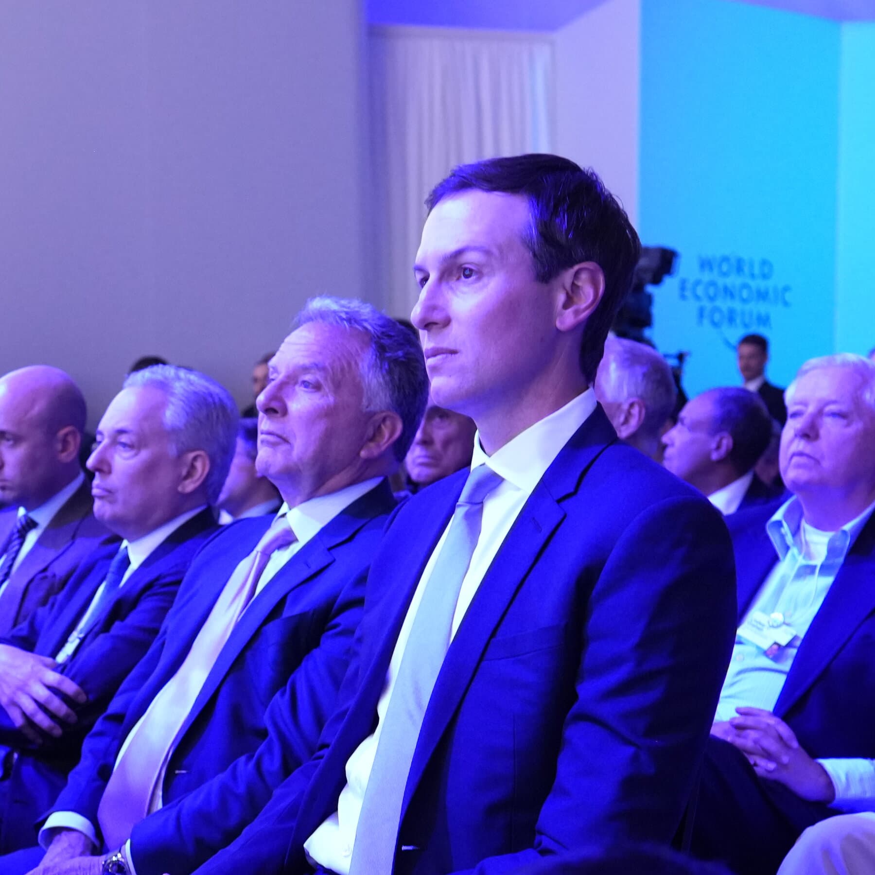 A hyper-realistic, cynical editorial illustration showing a sleek, polished conference table in Geneva. On one side sits a pristine document titled 'Guiding Principles' with nothing written on it. On the other side sits a cartoonishly large, lit stick of dynamite. In the background, blurry figures in suits shake hands, ignoring the explosive on the table. The lighting is cold and clinical, emphasizing the sterility of the room against the danger of the situation.