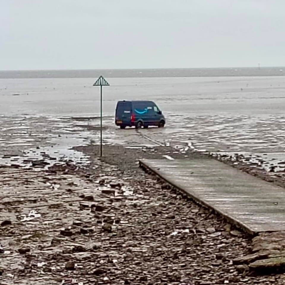 A white Amazon delivery van stuck deep in dark grey mud flats with the ocean tide rising around the wheels, gloomy overcast sky, ancient wooden marker posts in the distance, realistic style.