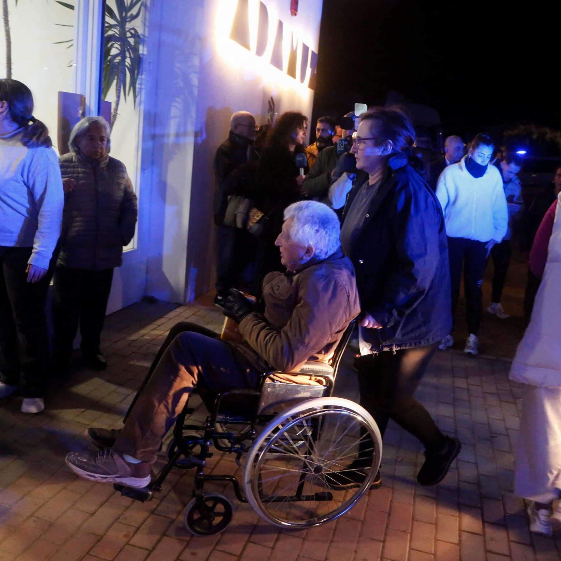A hyper-realistic, desaturated editorial illustration of a sleek, modern high-speed train violently twisting off the tracks in a dry, sun-baked Spanish landscape. The metal of the train is warping like fluid. In the foreground, out of focus, are the blurry backs of politicians in suits observing the scene with indifference. The lighting is harsh midday sun, casting long, sharp shadows. The mood is cold, sterile, and catastrophic.