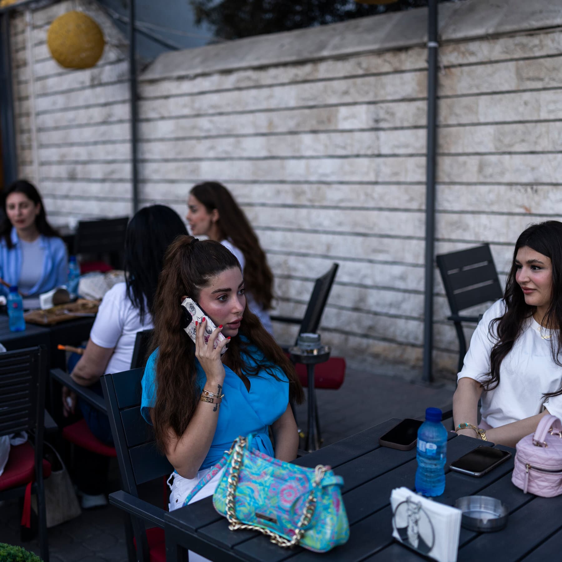 A hyper-realistic, gritty satirical illustration of a gray, crumbling government office in Syria. In the foreground, a large, absurd warning sign with a red circle and slash over a lipstick tube and a mascara wand. Behind the sign, tired female office workers in modest clothing look annoyed and exhausted, while a faceless male bureaucrat in a suit points an accusatory finger at them. The walls are cracked, and outside the window, there is vague ruin, contrasting the petty rule with the harsh reality.