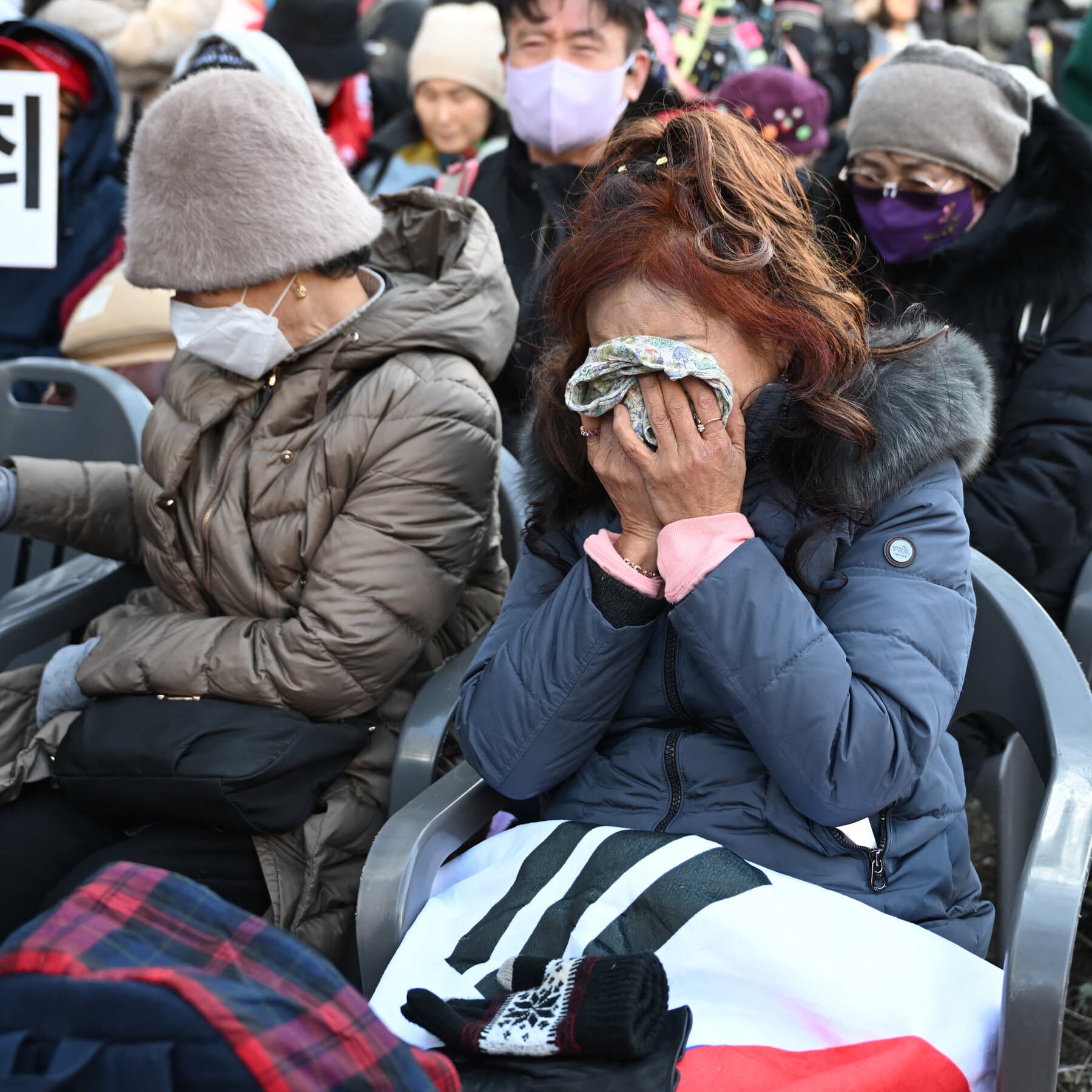 A split-screen composition showing a chaotic street scene in Seoul. On the left side, angry protesters with red signs shouting aggressively. On the right side, angry protesters with blue signs shouting aggressively. In the middle, a thick line of riot police with shields looking bored and tired. In the background, a gray, imposing courthouse building looms over the crowd. The sky is overcast and gloomy. The style should be gritty and realistic photojournalism.
