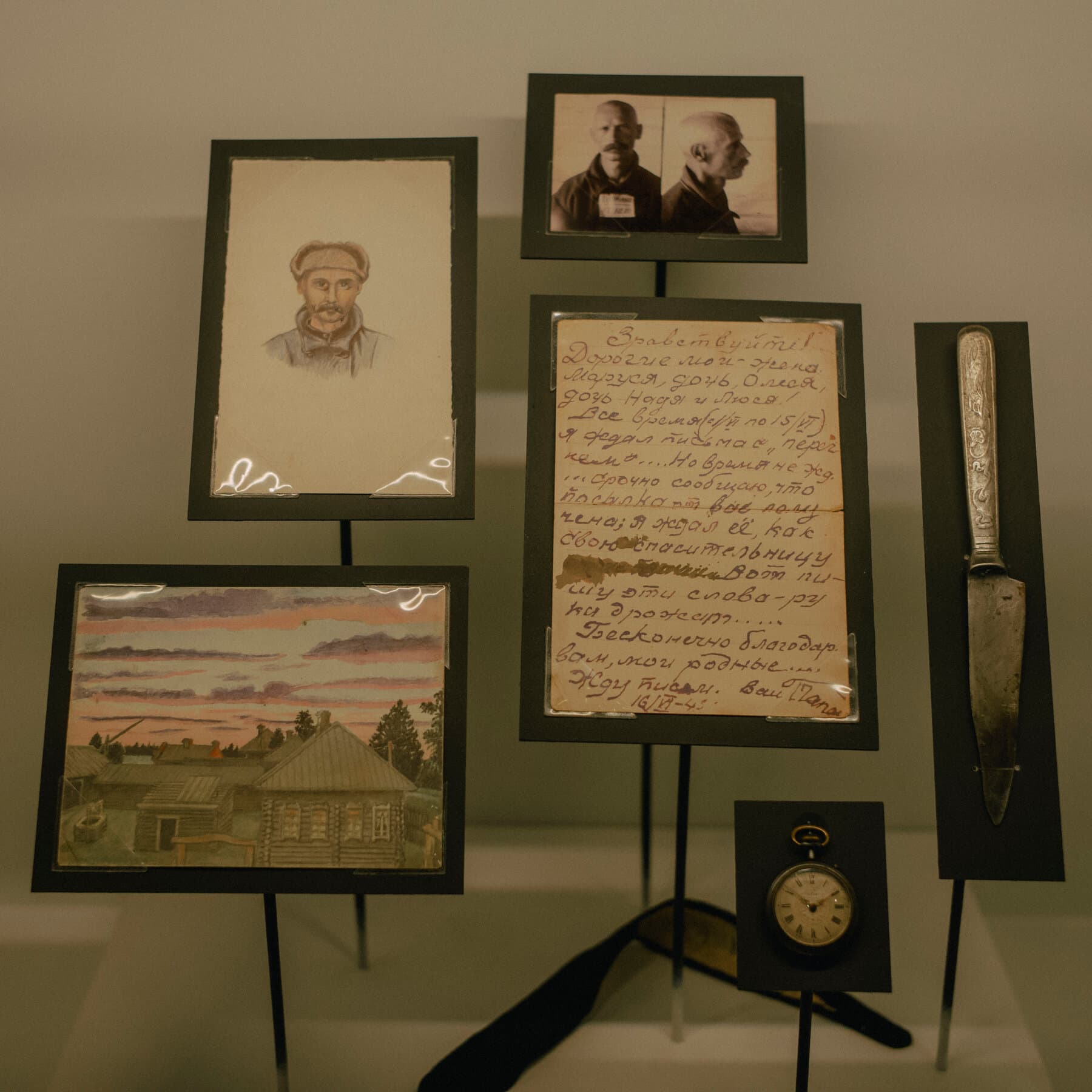 A stark, gloomy illustration of a museum hallway. The walls are being painted over with grey paint, covering up black-and-white photos of sad prisoners. In the center, a large, ornate rug is being pulled over a pile of human skulls. The lighting is dim and cold. Digital art style.
