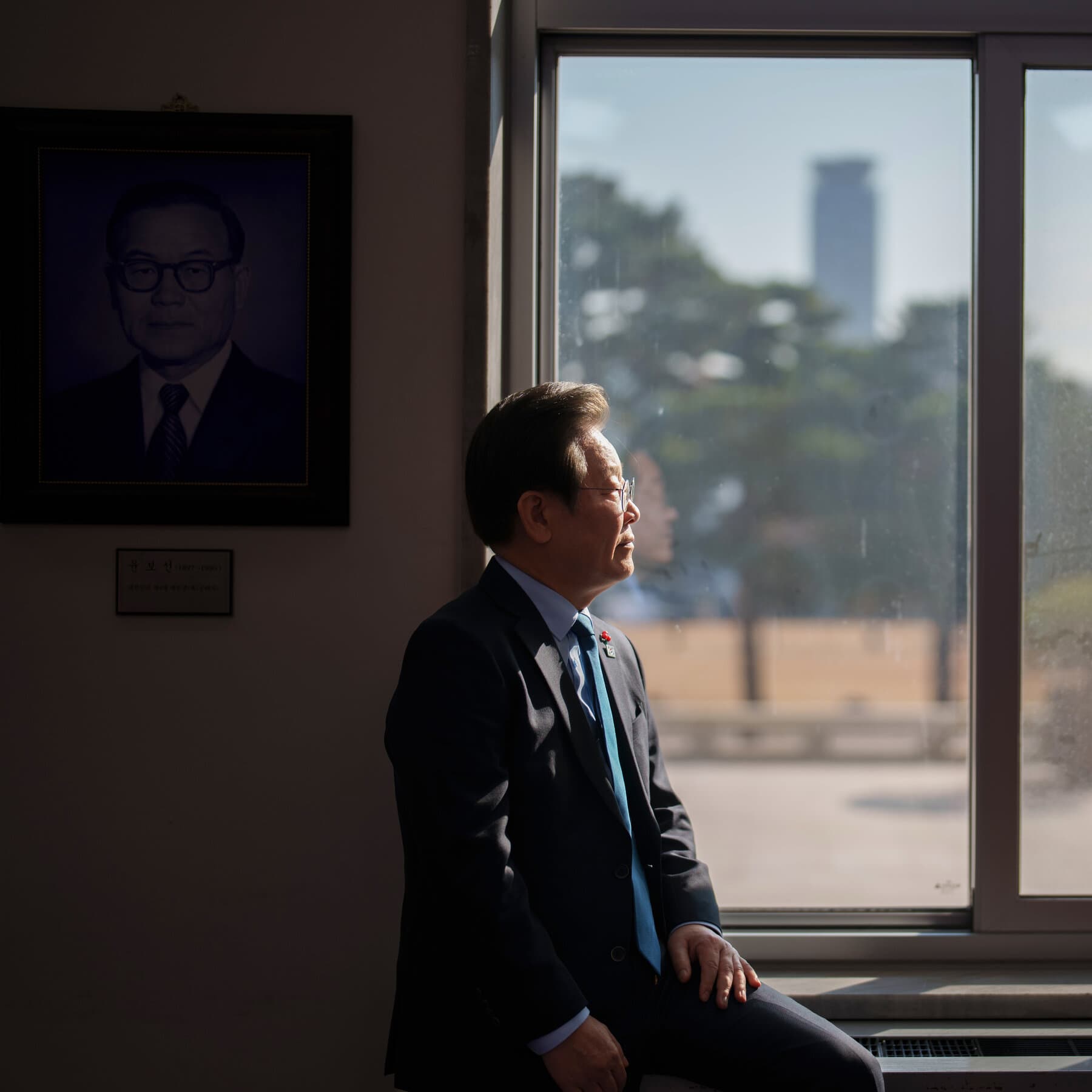 A high-contrast, noir-style black and white illustration of a lonely political figure standing on a crumbling concrete podium in Seoul. The figure is small compared to the massive, oppressive city skyline in the background. The scene is split down the middle by a jagged crack in the ground. The atmosphere is gloomy, cynical, and cold.